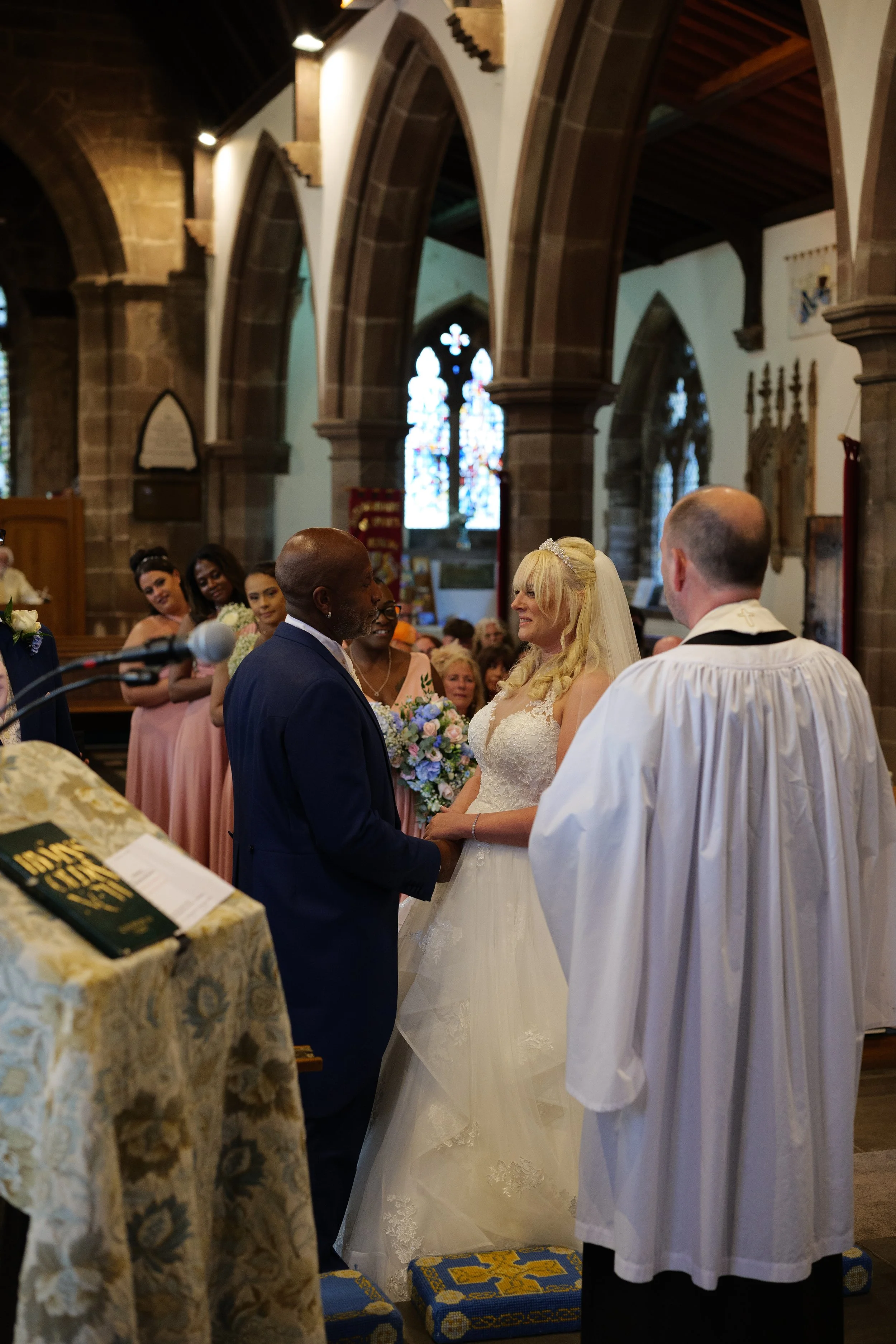 A wedding ceremony inside a church, with the bride and groom holding hands and facing each other, surrounded by bridesmaids and officiant.