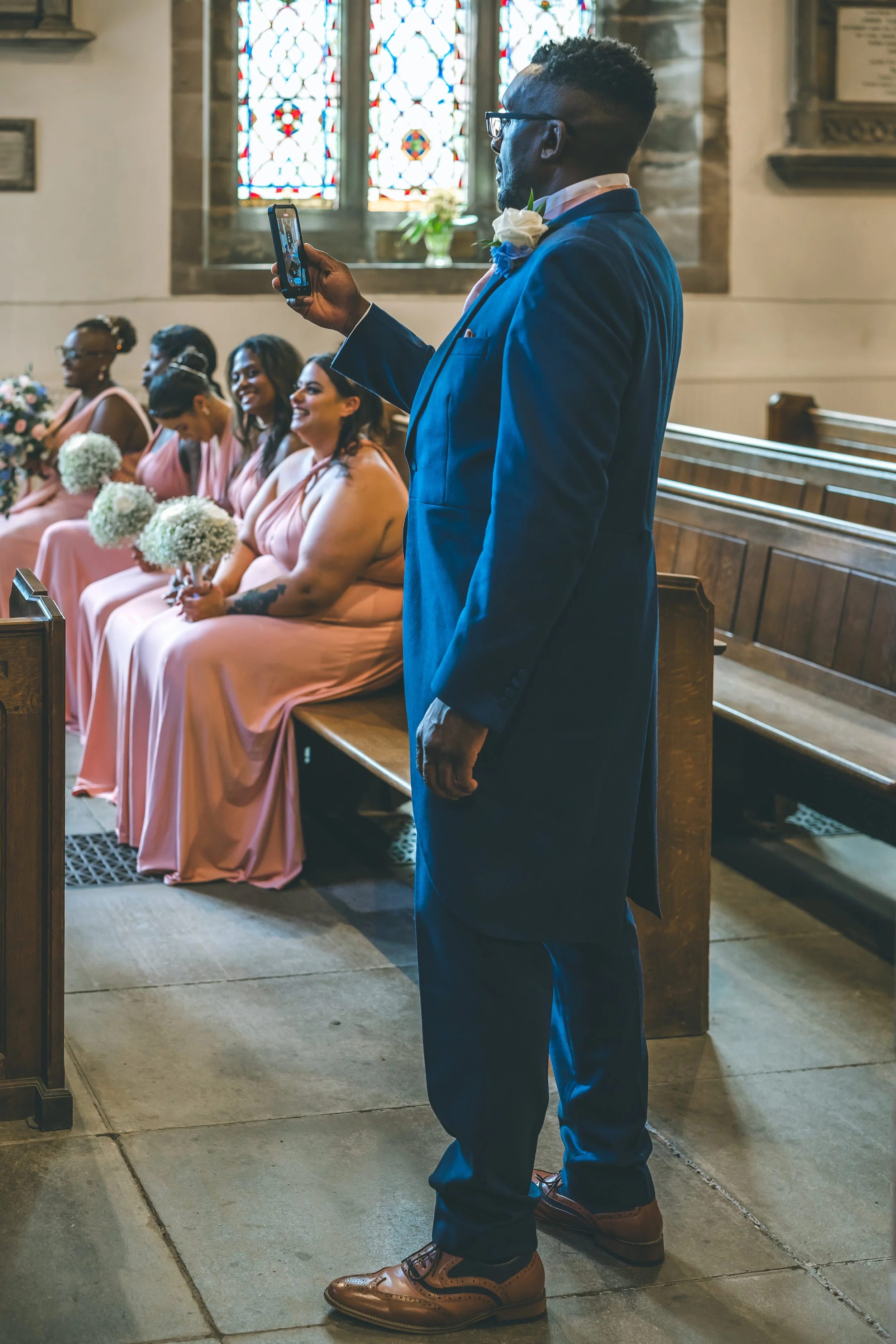 A man in a blue suit and brown shoes is taking a photo or video with his phone inside a church. Behind him, six women in matching pink dresses with white flowers are sitting in pews, holding bouquets, and smiling. The church has stained glass windows