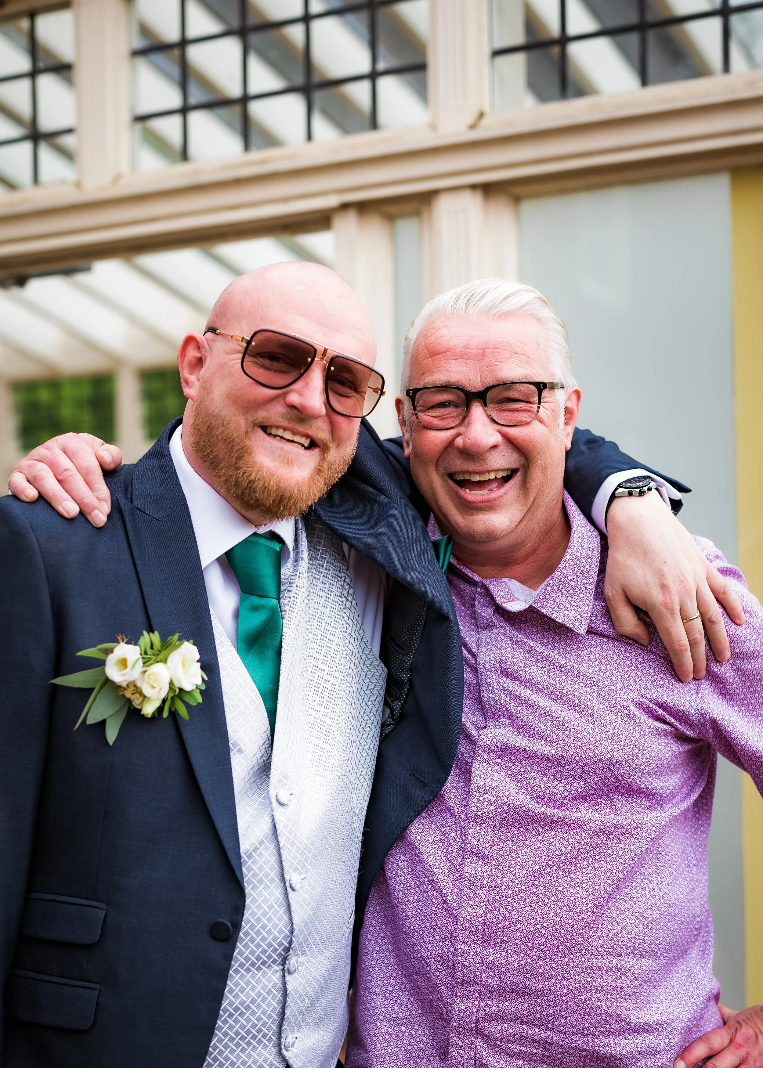 Two men with glasses smiling and hugging each other at an outdoor event, one wearing a suit with a boutonniere and the other in a patterned shirt, in front of a glass and wood structure.