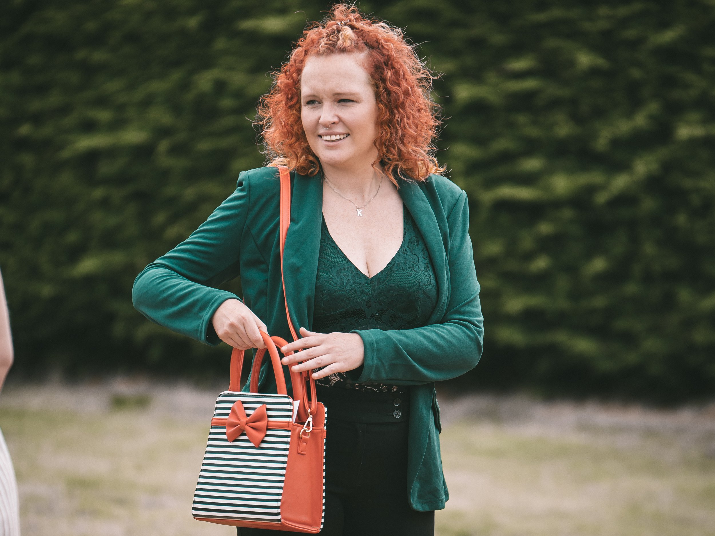 A woman with curly red hair and light skin is smiling and looking to her left. She is wearing a green blazer over a dark green lace top, with a necklace that has a small letter 'K' pendant. She is outdoors with blurred green foliage in the background