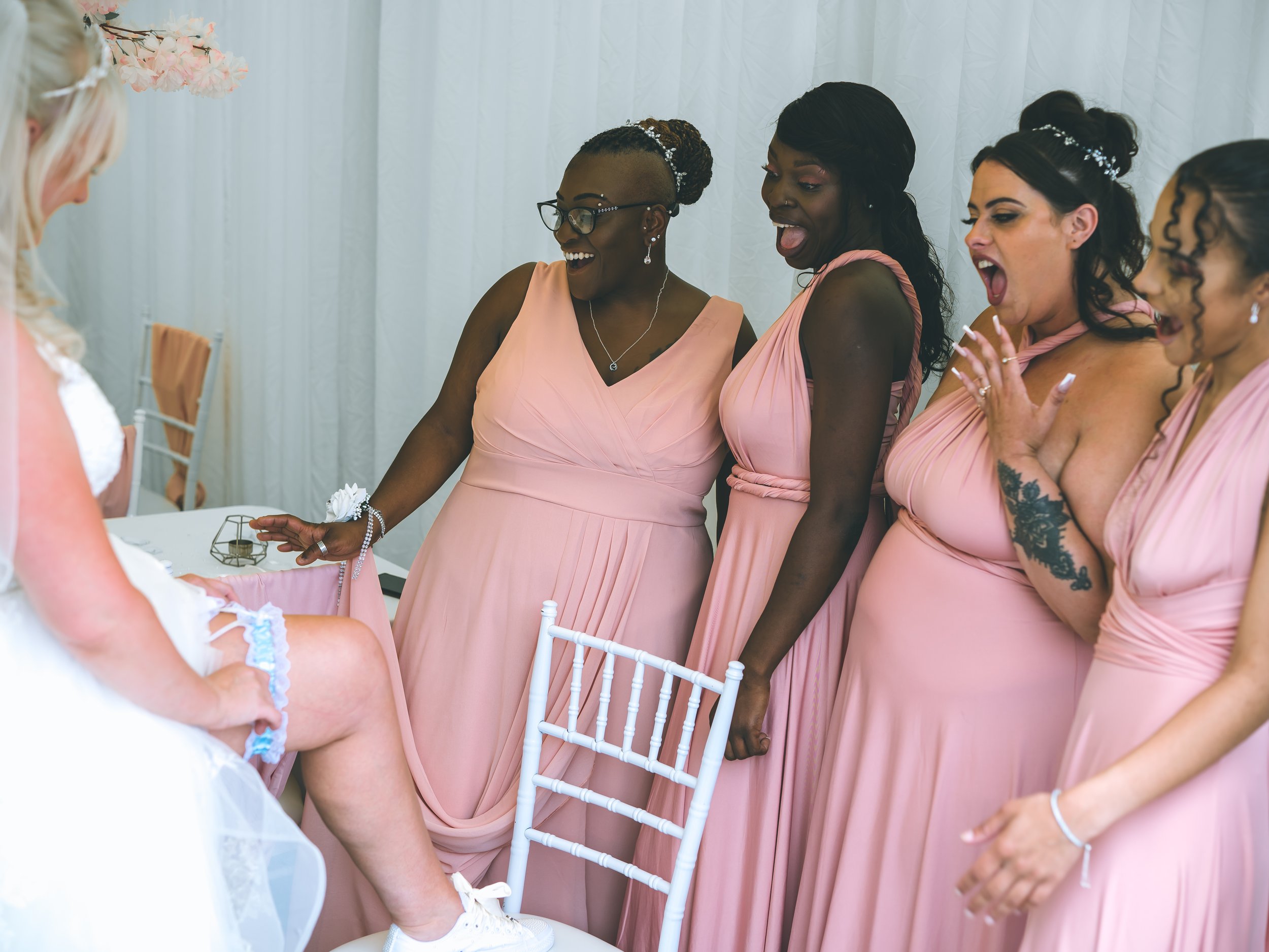 A group of women dressed in pink bridesmaid dresses gathered around a bride during a wedding celebration, smiling and reacting happily.