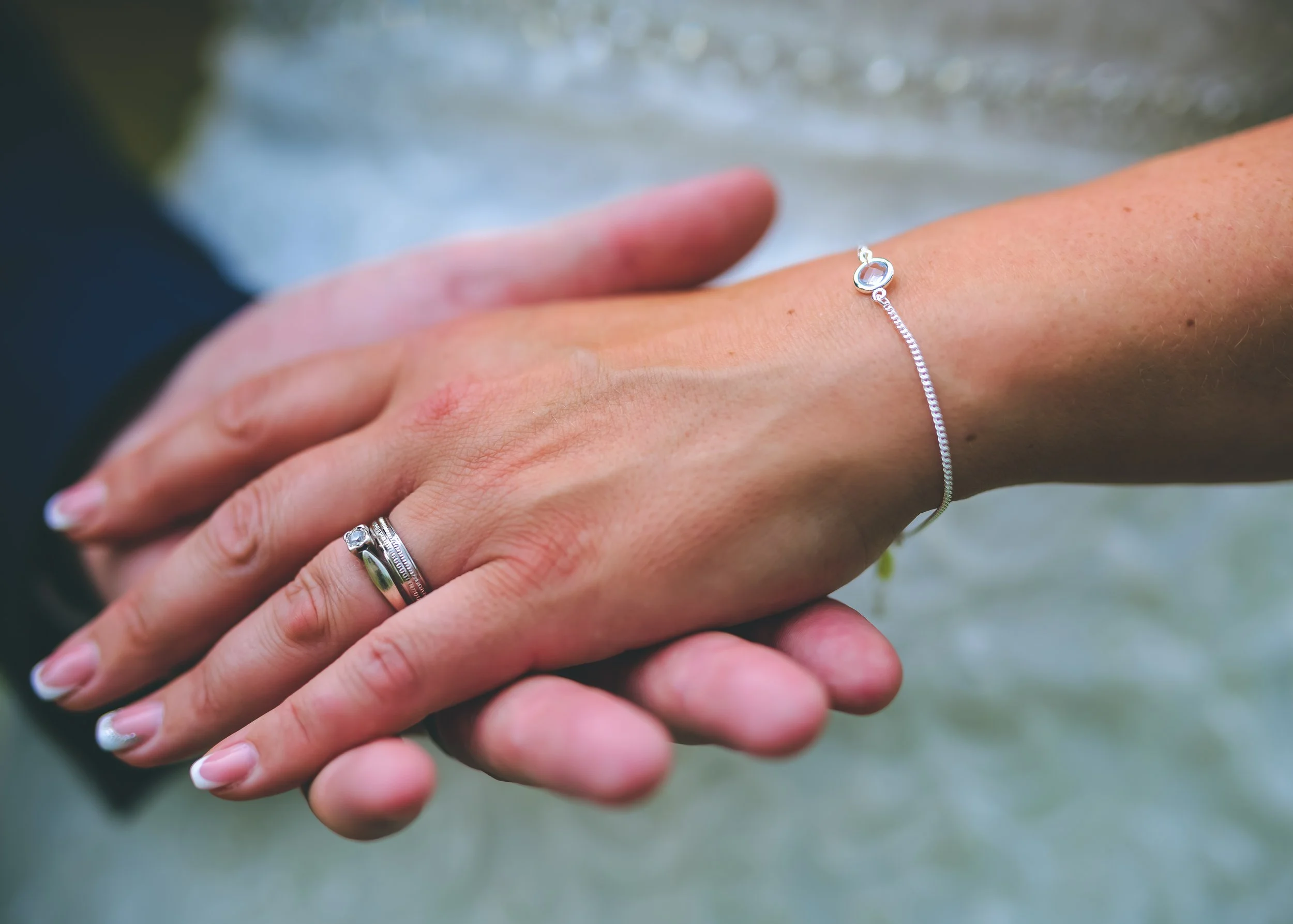 Close-up of a person's hand with a wedding ring, holding another hand with a silver bracelet and ring, outdoors.