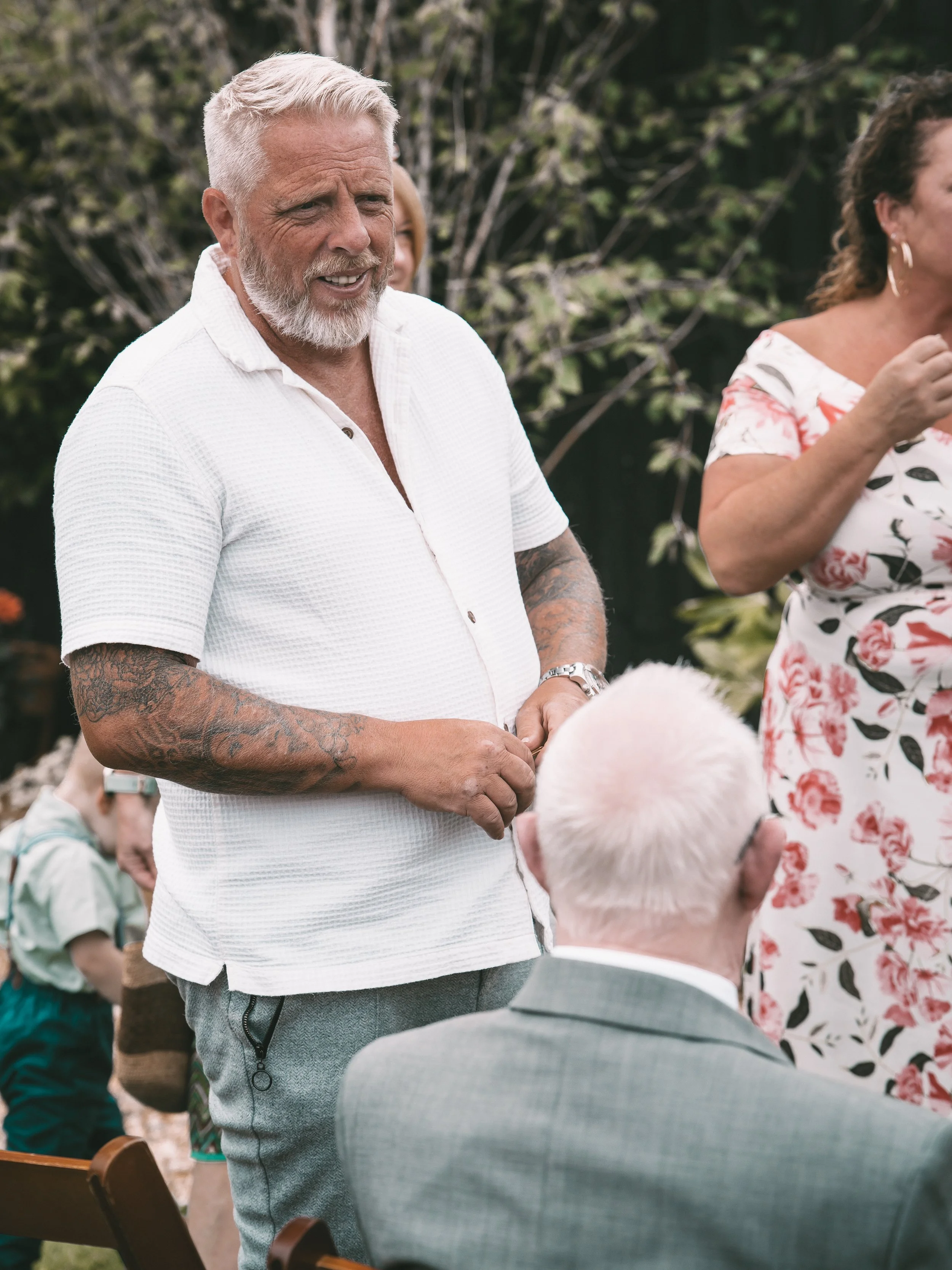An older man with a white beard and tattoos on his arms, wearing a white shirt, standing outdoors and smiling, while people around him are engaged in conversation.