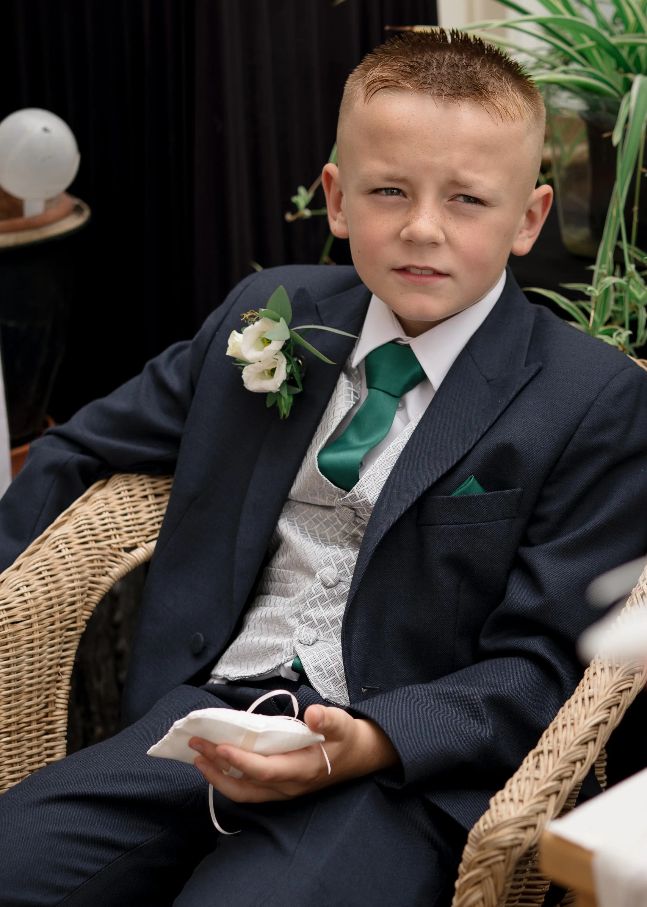 A young boy with short, light brown hair, wearing a navy blue suit with a white dress shirt, green tie, and a white boutonniere, sitting in a wicker chair at a formal event, holding a face mask in his hand.