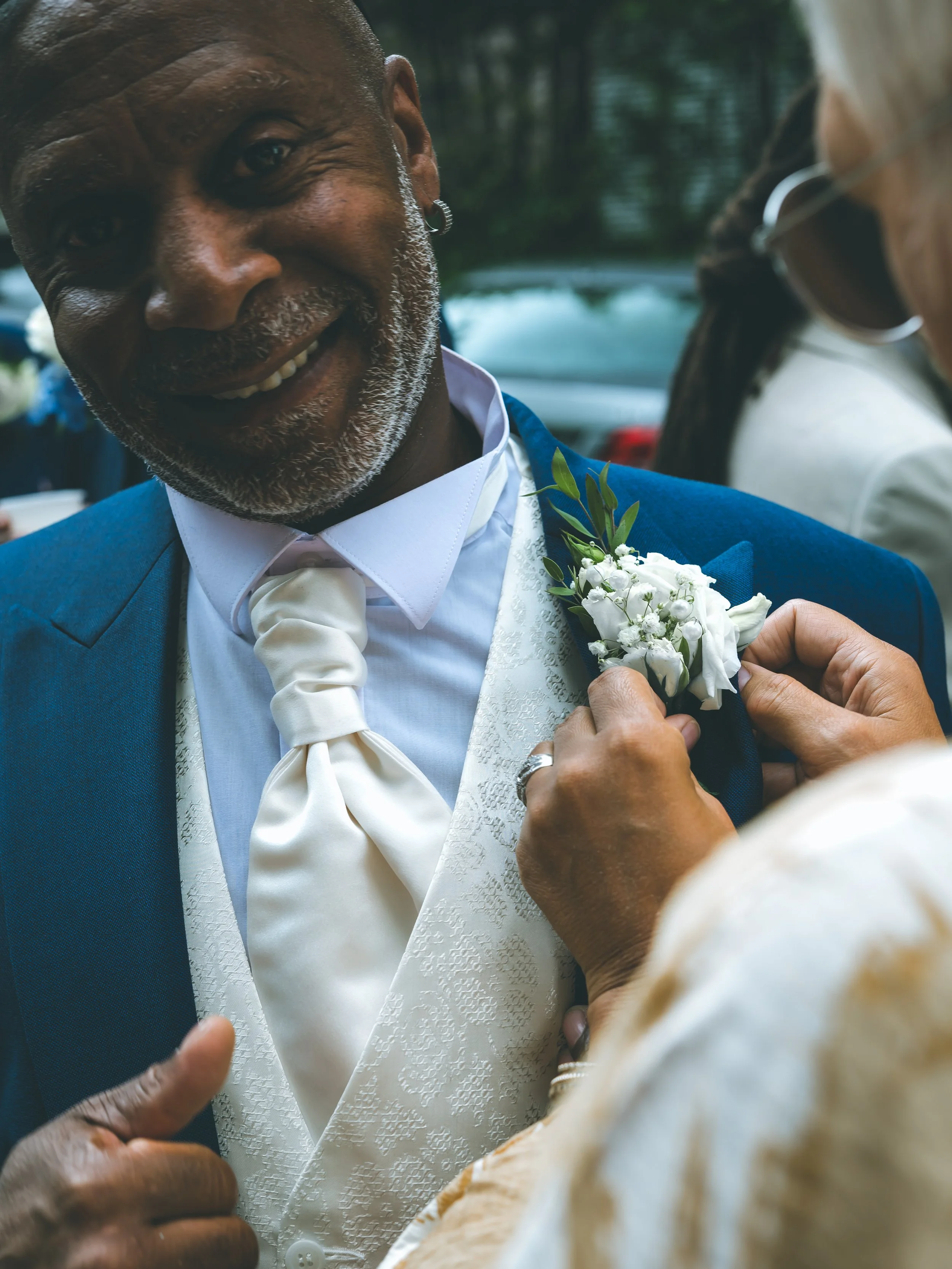 A man in a blue suit with a white shirt and tie receives a boutonniere from someone at an outdoor event.
