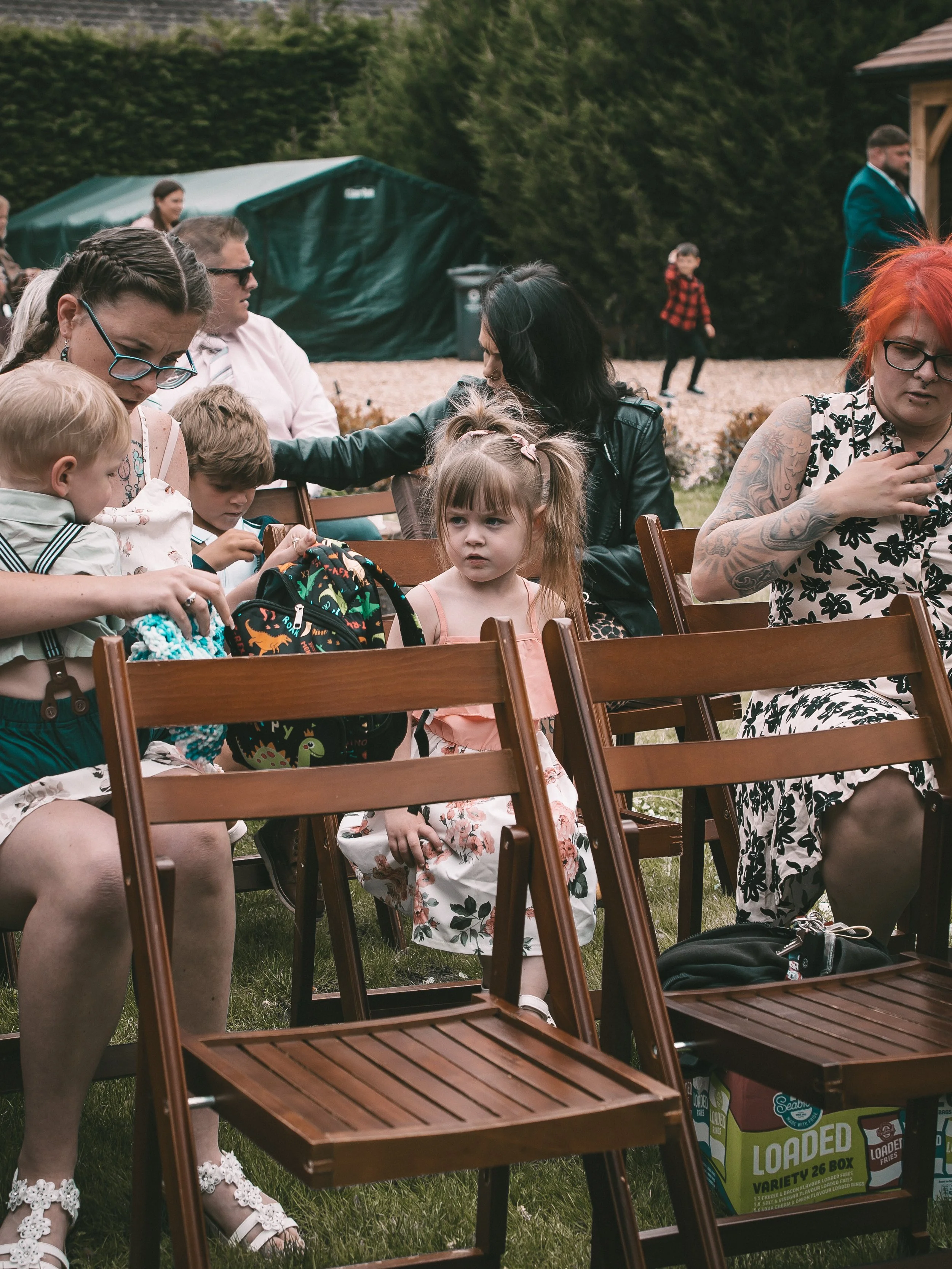A group of children and adults sitting outdoors on wooden chairs, with some children looking at objects in their hands. In the background, there are trees, a green tent, and a man in a teal blazer.