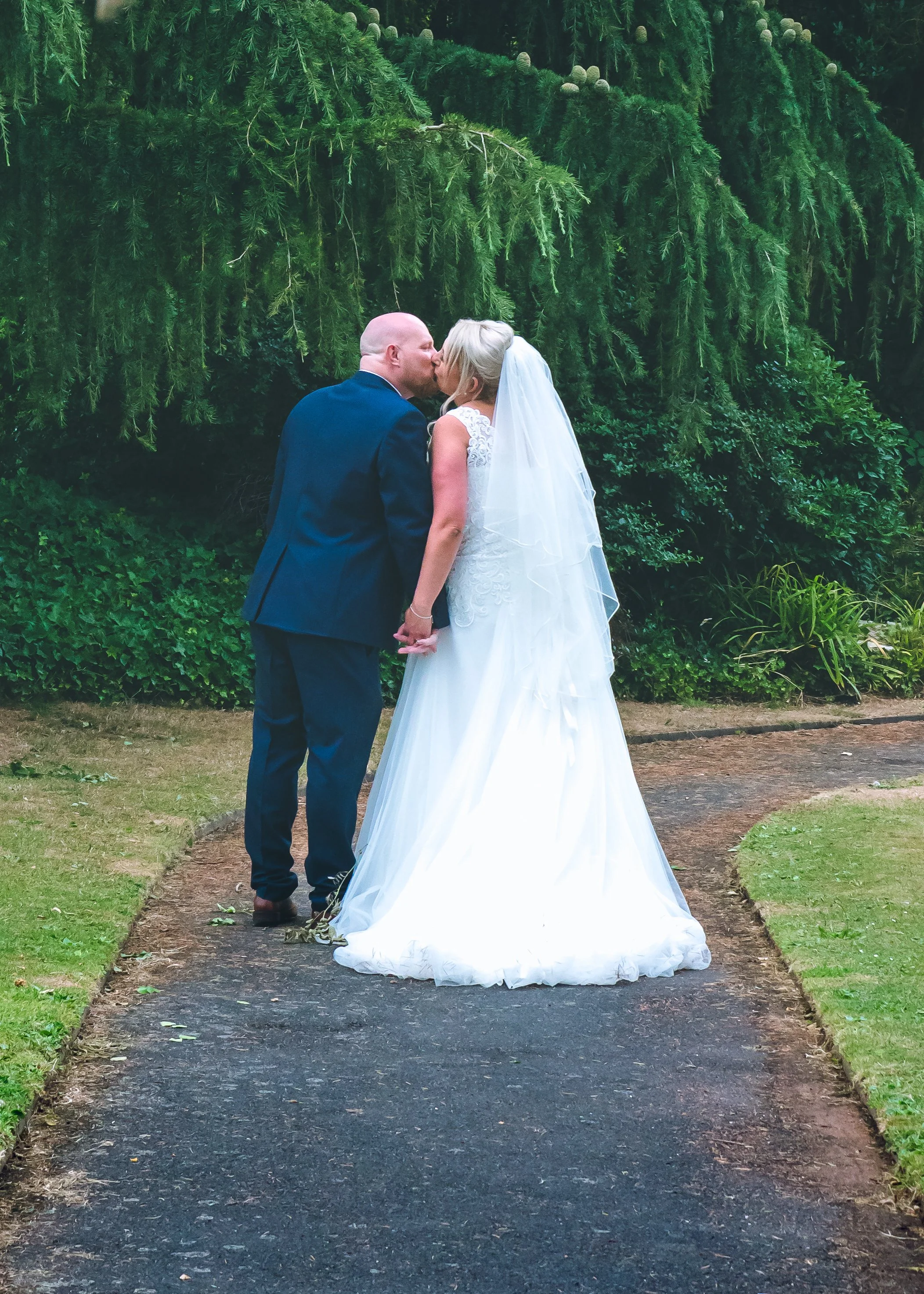 A bride and groom share a kiss outdoors on a dirt path surrounded by lush greenery, with the bride wearing a white wedding dress and veil, and the groom in a dark suit.