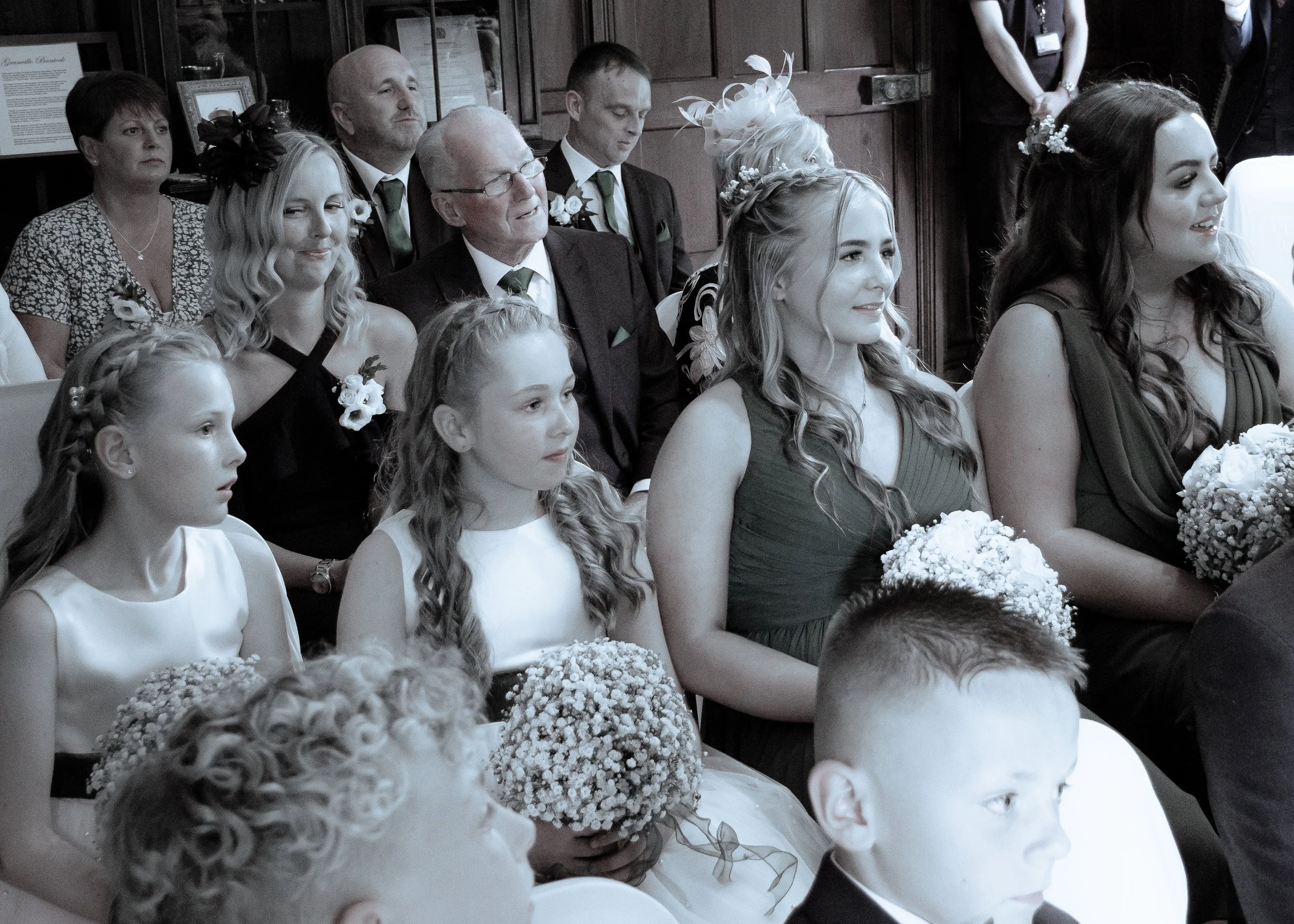 A group of people attending a wedding ceremony, including young girls in white dresses holding bouquets, and adults in formal attire, seated inside a church or similar venue.