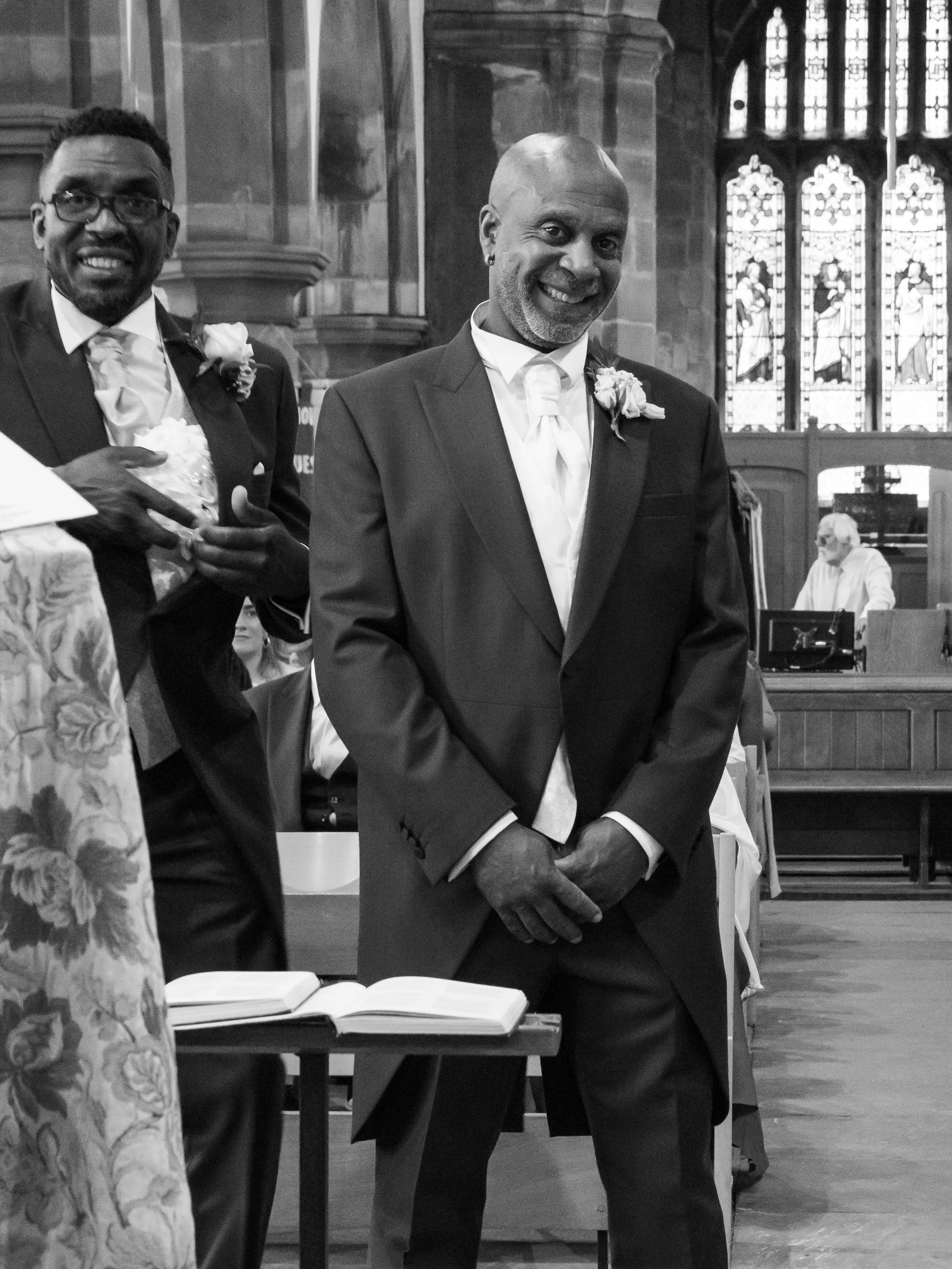 A black and white photo of a man in a dark suit, white shirt, and tie, standing inside a church with stained glass windows, with a person seated at a keyboard in the background.
