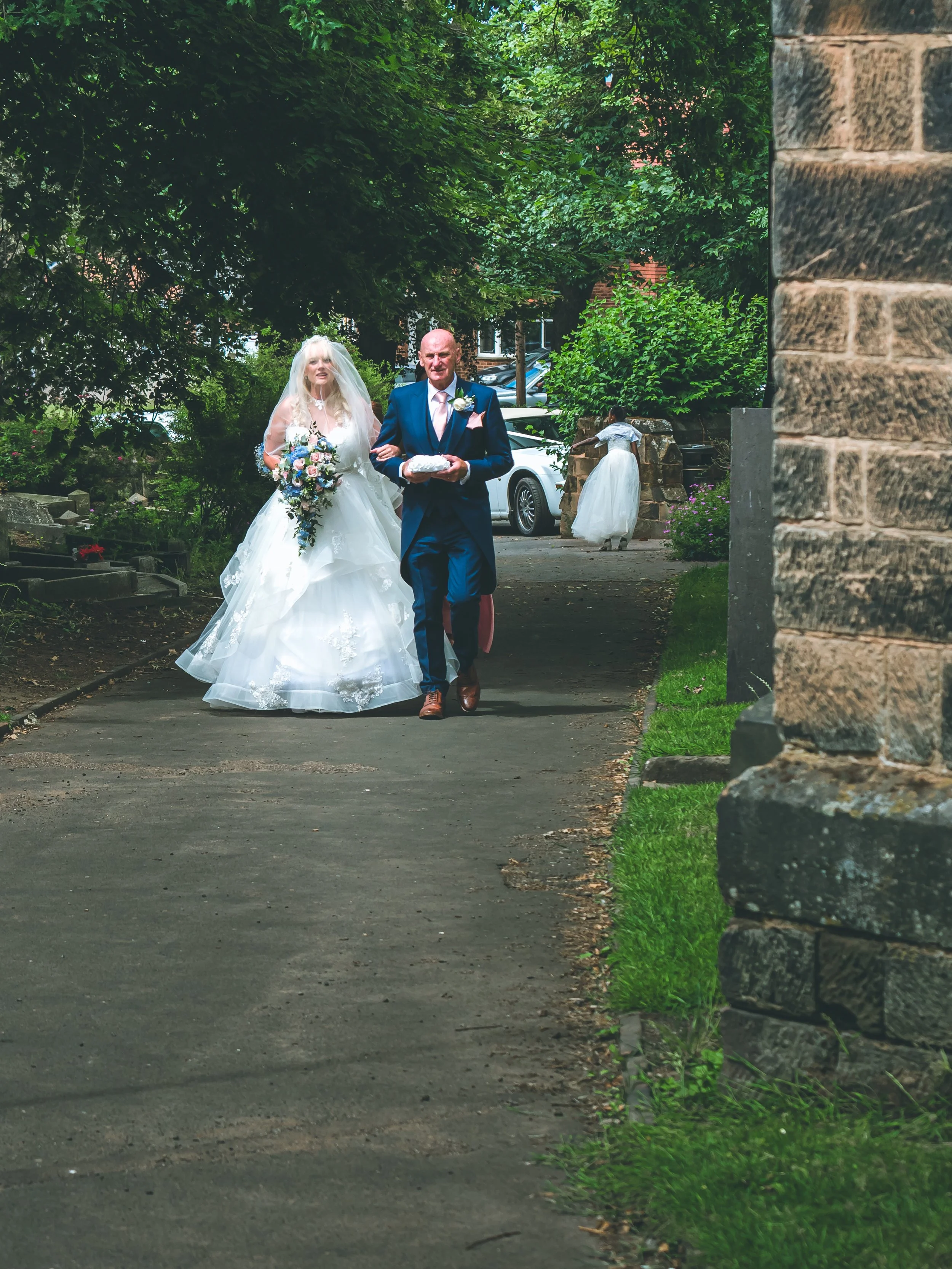 A bride and groom walking outdoors on a paved path, surrounded by trees and greenery, with the bride holding a large bouquet of flowers and the groom holding a wrapped item while an assistant or helper stands in the background.