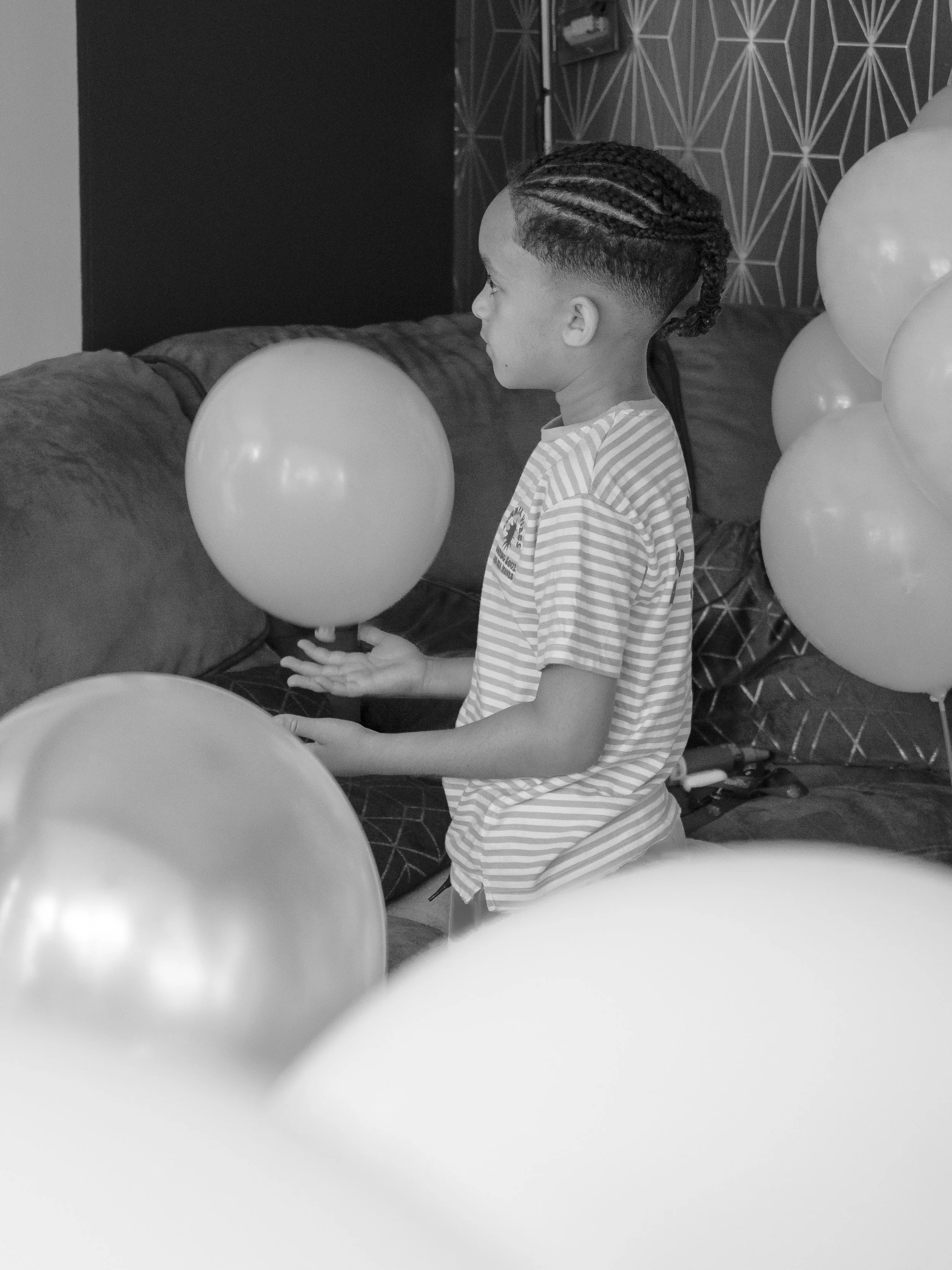 A young boy with braided hair, wearing a striped shirt, standing near a cluster of balloons, in a room with patterned wallpaper.