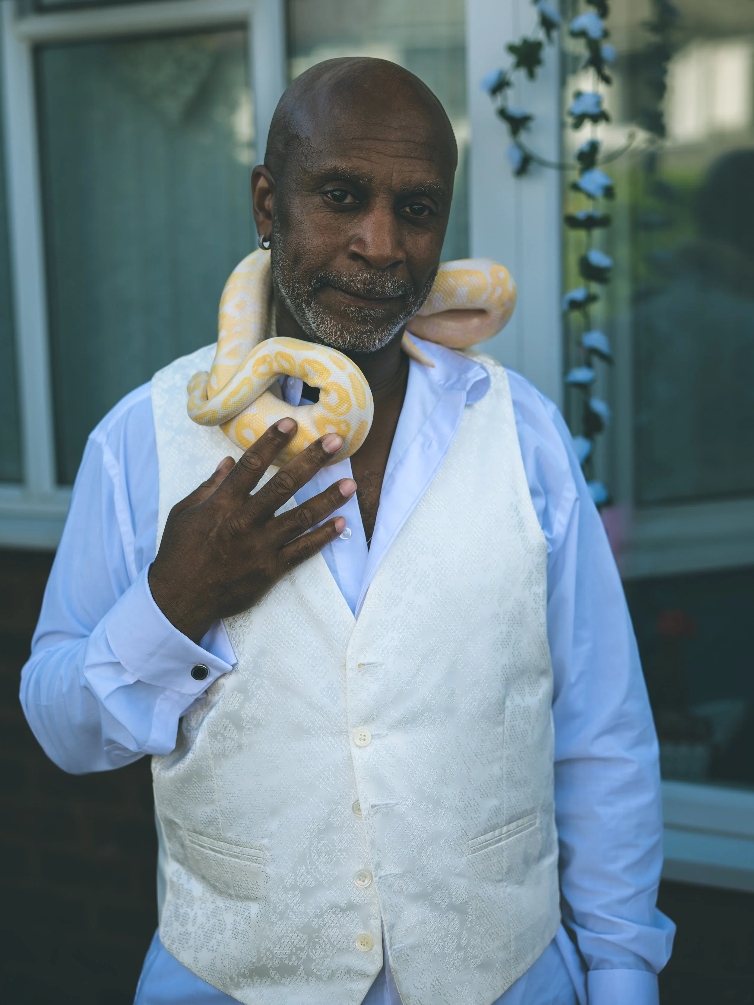 An older man with a bald head, gray beard, and earring, wearing a white shirt and cream-colored vest, holding a yellow and white ball python around his neck, standing outdoors near a glass building with a plant in the background.