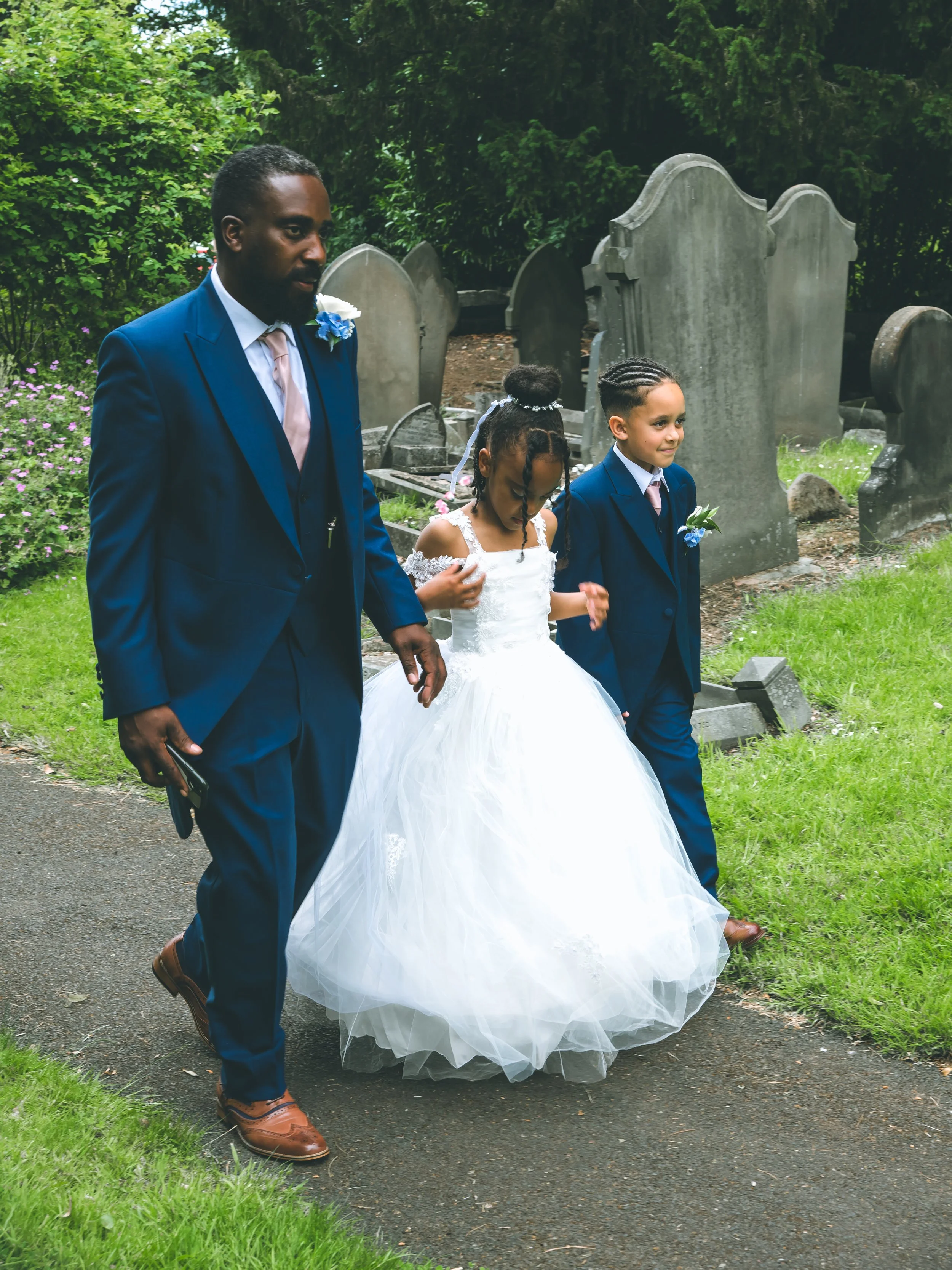 A man, woman, and two children walking in a cemetery. The woman is in a white wedding dress, and the man and boy are in blue suits with pink ties. The girl is in a white dress with lace, and the setting is outdoor with green grass and trees.