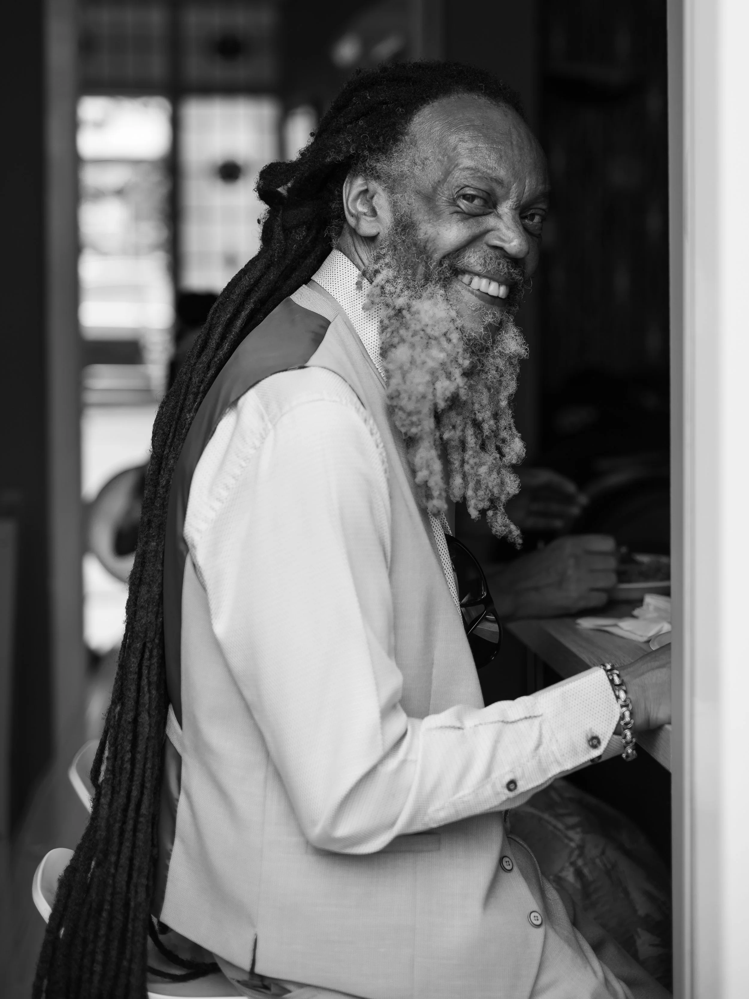 A smiling elderly man with long dreadlocks and a beard, wearing a light-colored blazer and patterned shirt, standing at a voting kiosk.