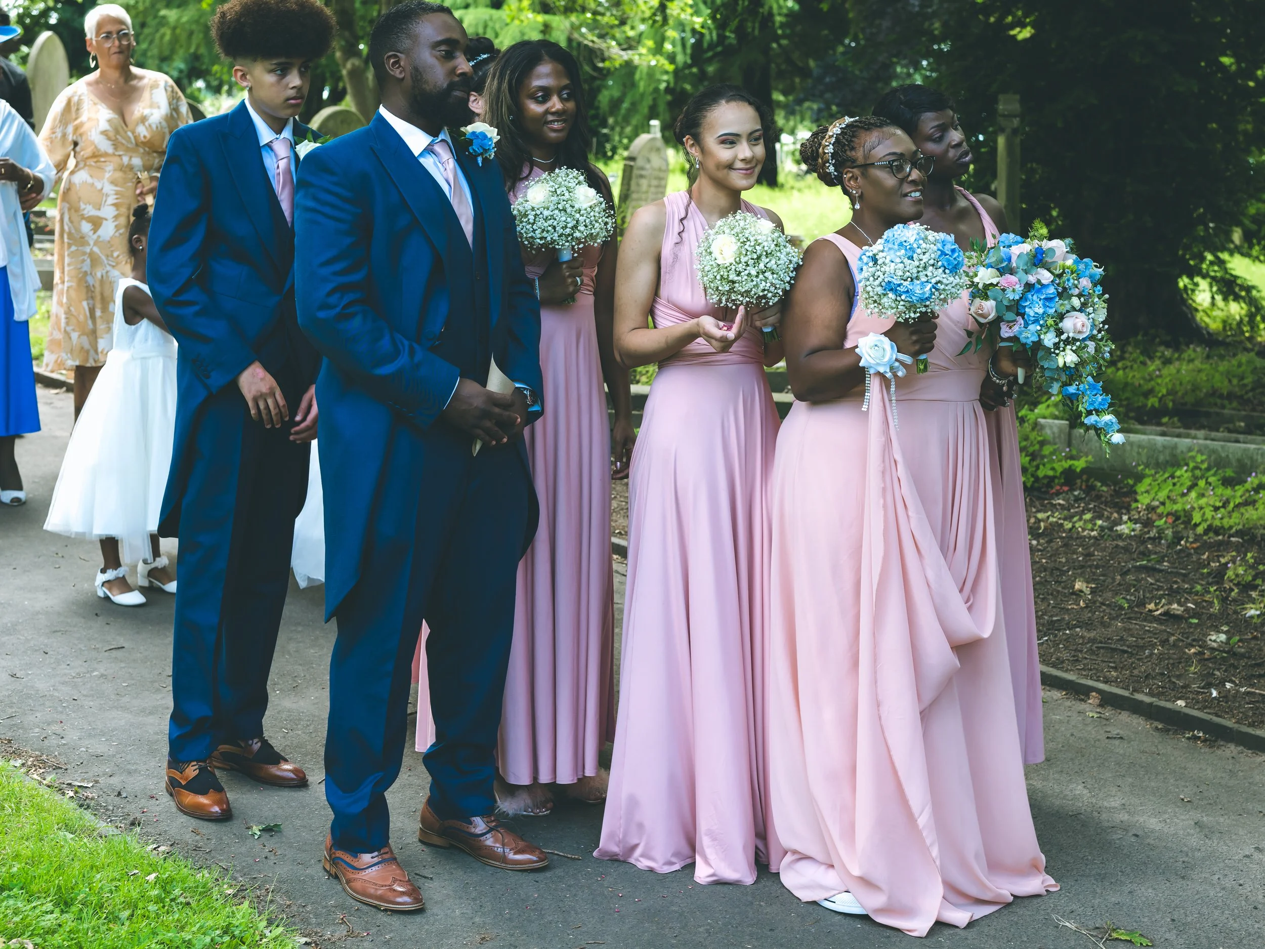 Group of bridesmaids and groomsmen standing outdoors on a pathway, dressed in formal attire, holding bouquets of blue and white flowers during a wedding ceremony in a lush green park.