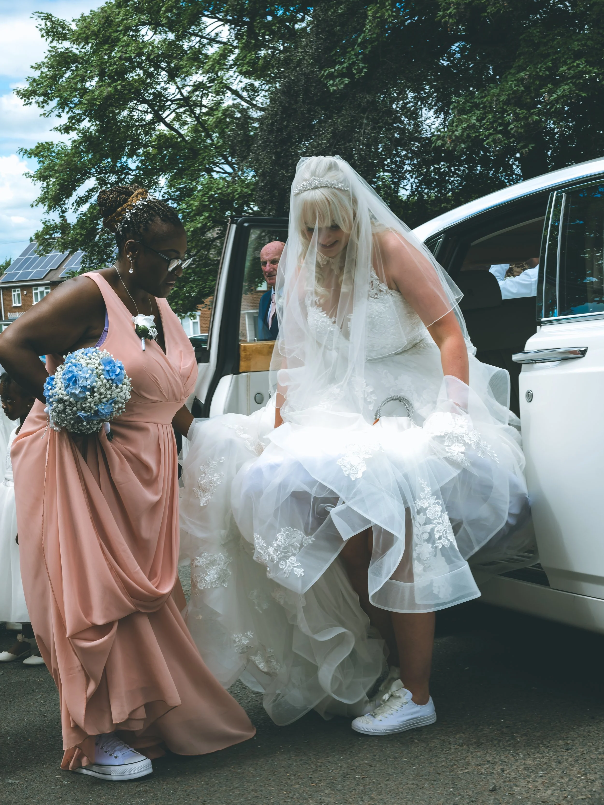 A bride in a wedding dress bending down, wearing white sneakers. A woman in a pink dress holding a bouquet of blue and white flowers stands nearby. A car trunk is open, with a man in a suit visible inside. Green trees and houses with solar panels are