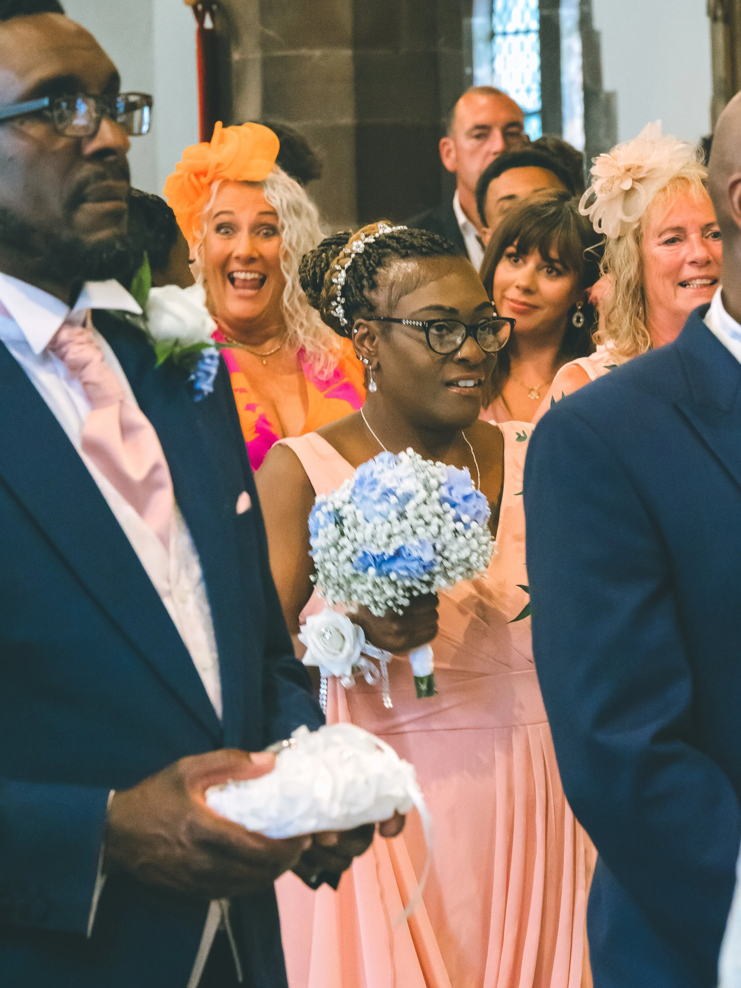 A group of diverse people gathered at a wedding ceremony, with a woman in the center holding a bouquet of blue and white flowers, wearing glasses and a pink dress.