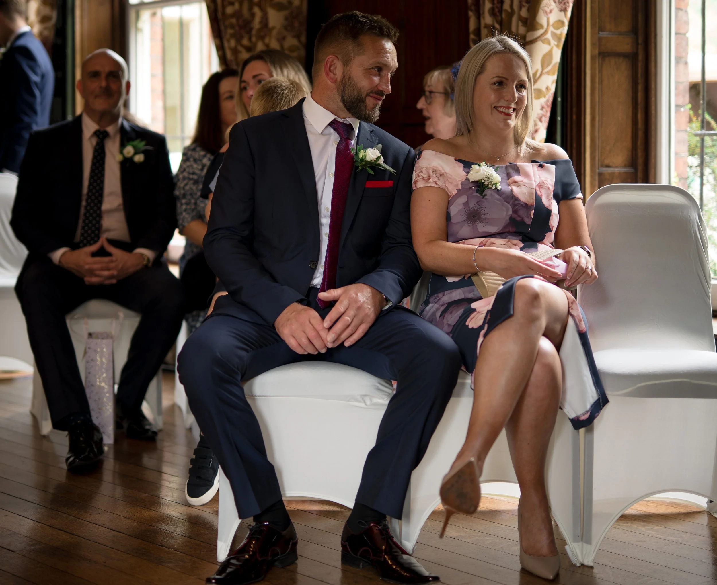 People sitting in chairs at a wedding ceremony, dressed formally, with a man in tuxedo and woman in floral dress in the foreground, smiling.