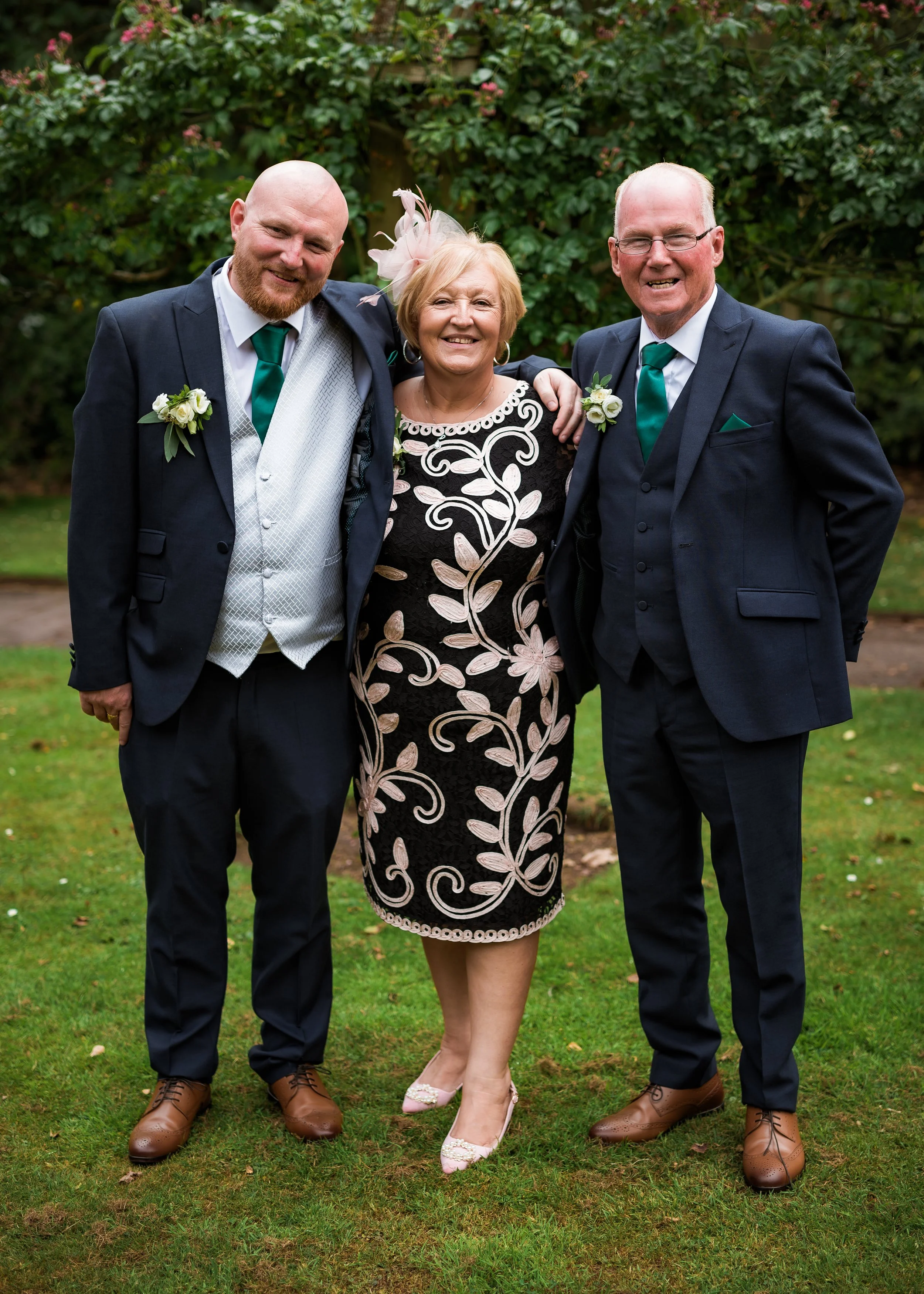 Three people standing outdoors on grass, smiling, dressed in formal wedding attire. The person in the middle is a woman wearing a black dress with white embroidered floral designs, pink heels, and a pink fascinator. The two men on either side are wea