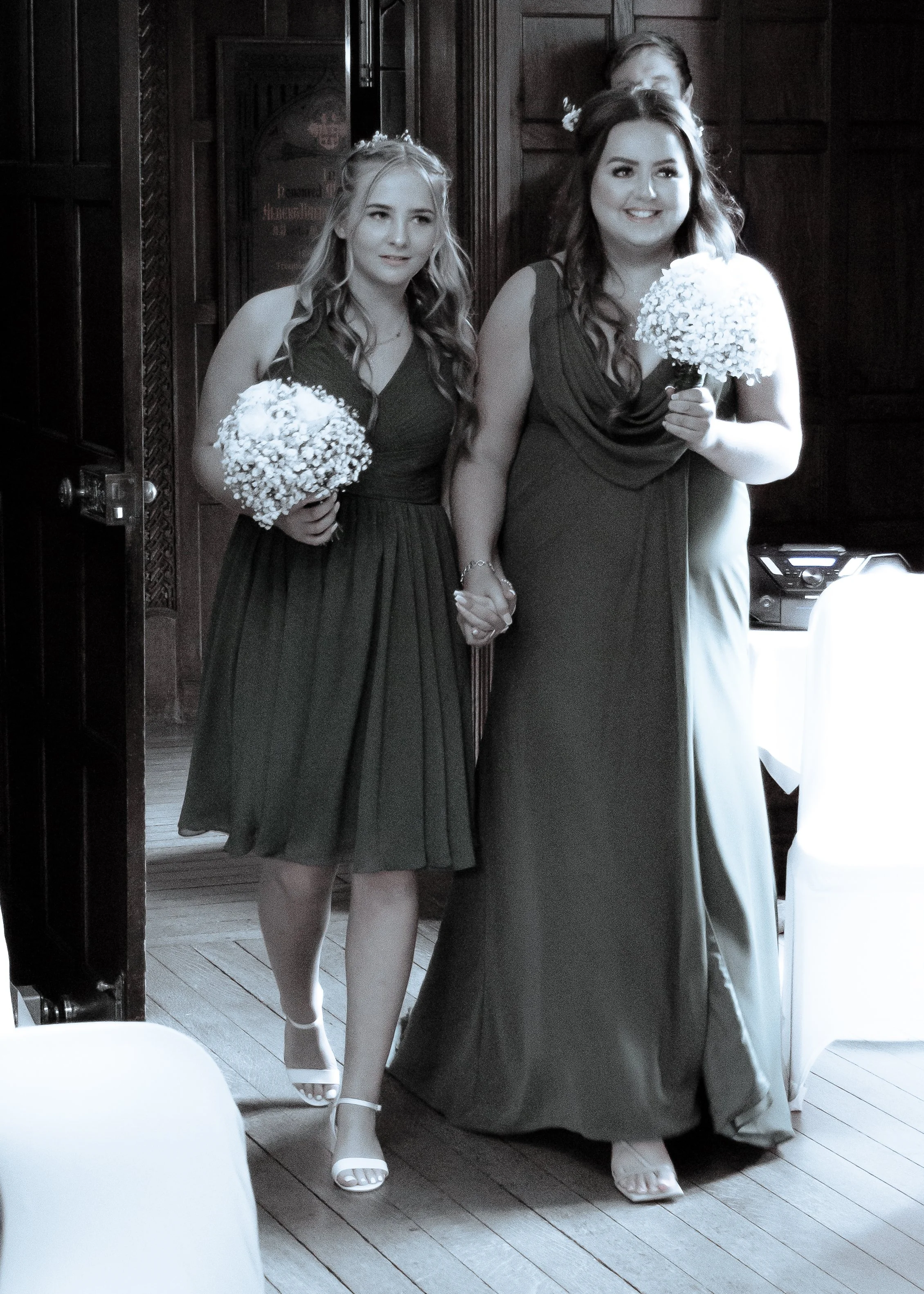 Two women holding hands and holding white floral bouquets, dressed in formal gowns at a wedding or special event in a wood-paneled room.