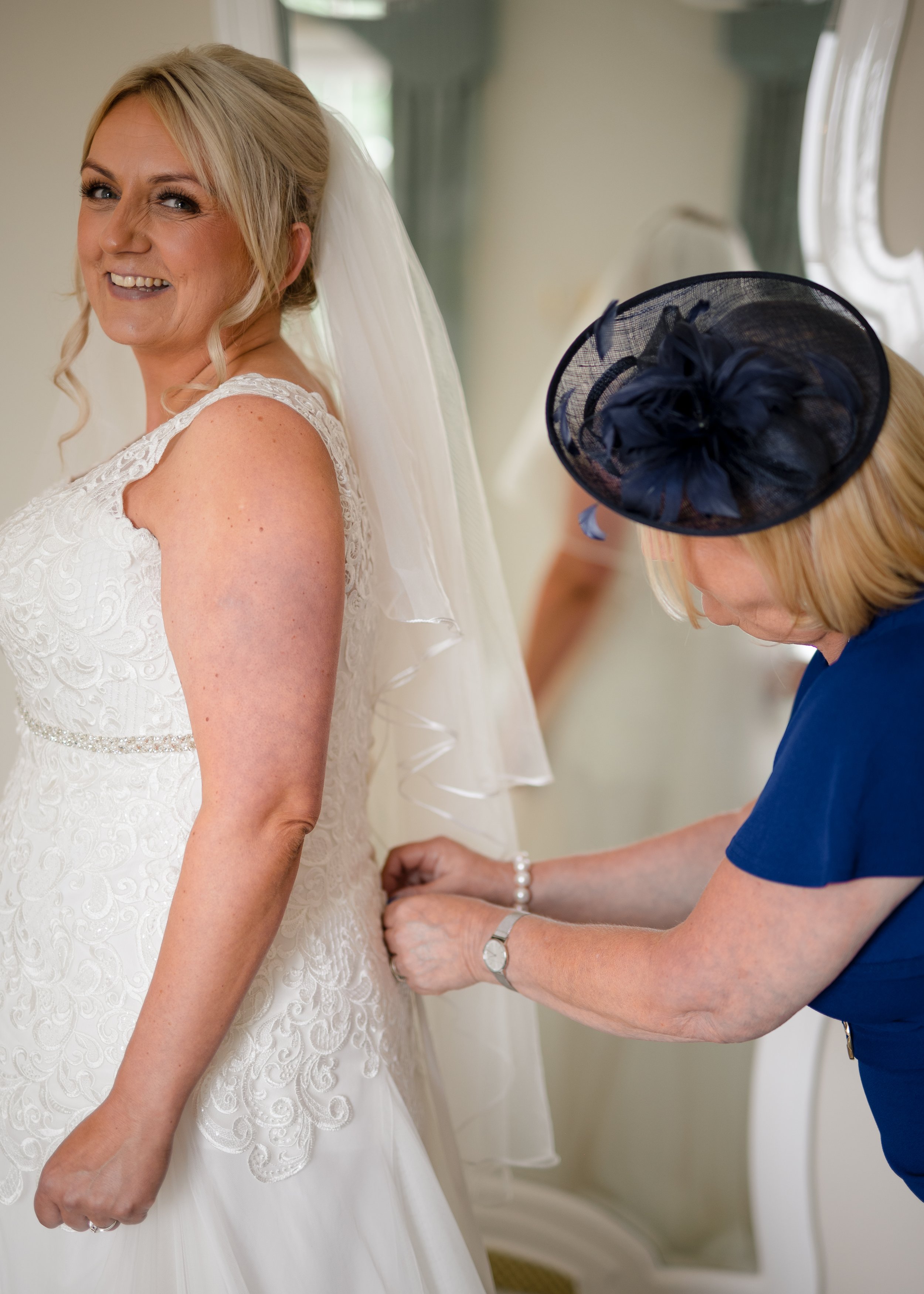 A smiling woman in a white wedding dress is having her dress fastened by another woman in a blue dress and a navy hat with a bow. The bride has blonde hair and is looking at the camera, standing in front of a mirror.