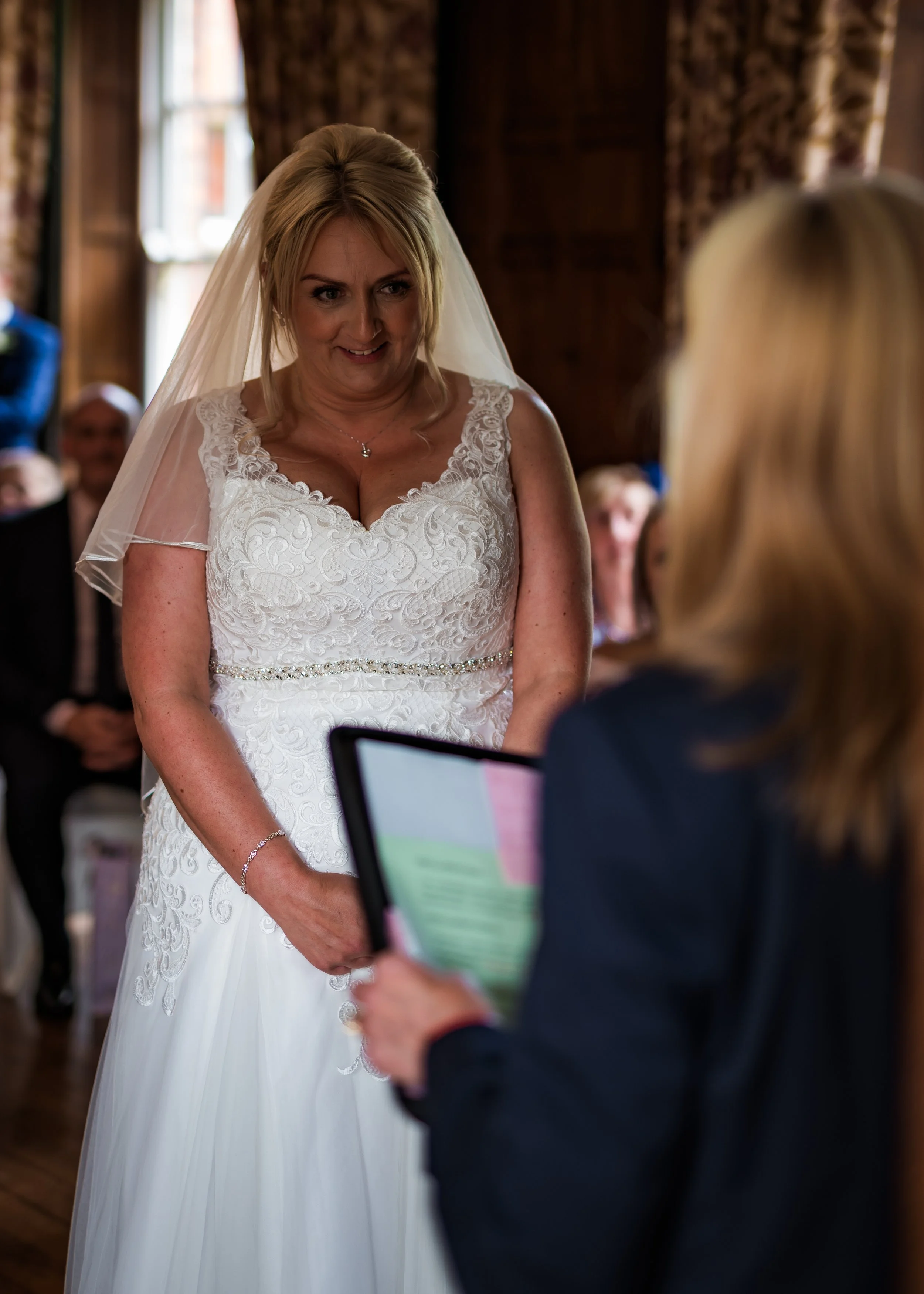 A bride with blonde hair in a white lace wedding dress and veil smiles as she looks at her partner during a wedding ceremony inside a wooden venue with curtains and large windows.