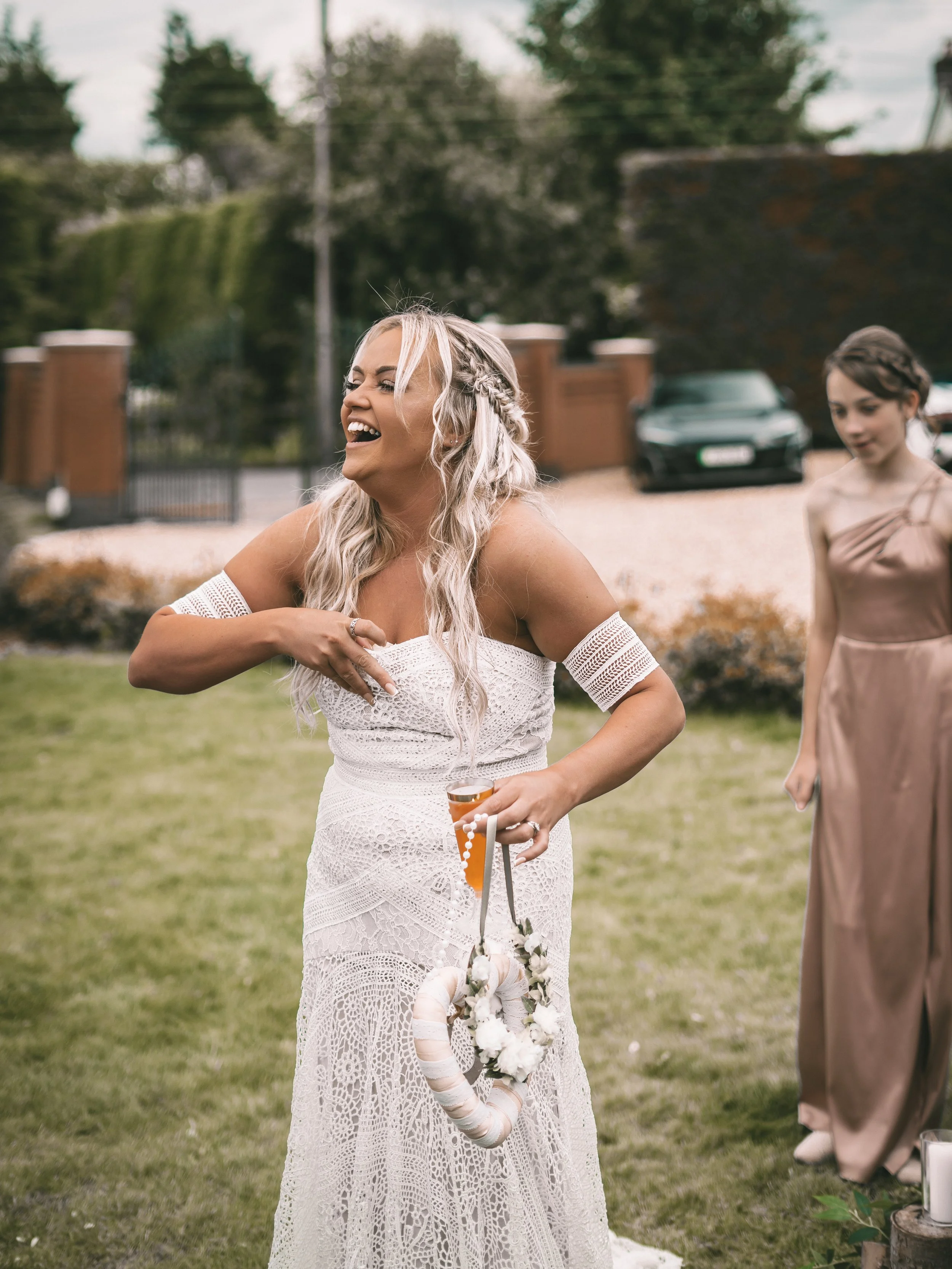 A woman with long, wavy blonde hair in a white lace dress laughing outdoors during a sunny day, holding a floral wreath and a small accessory in her hand. Another woman in a brown dress is visible in the background, standing on grass with trees and a