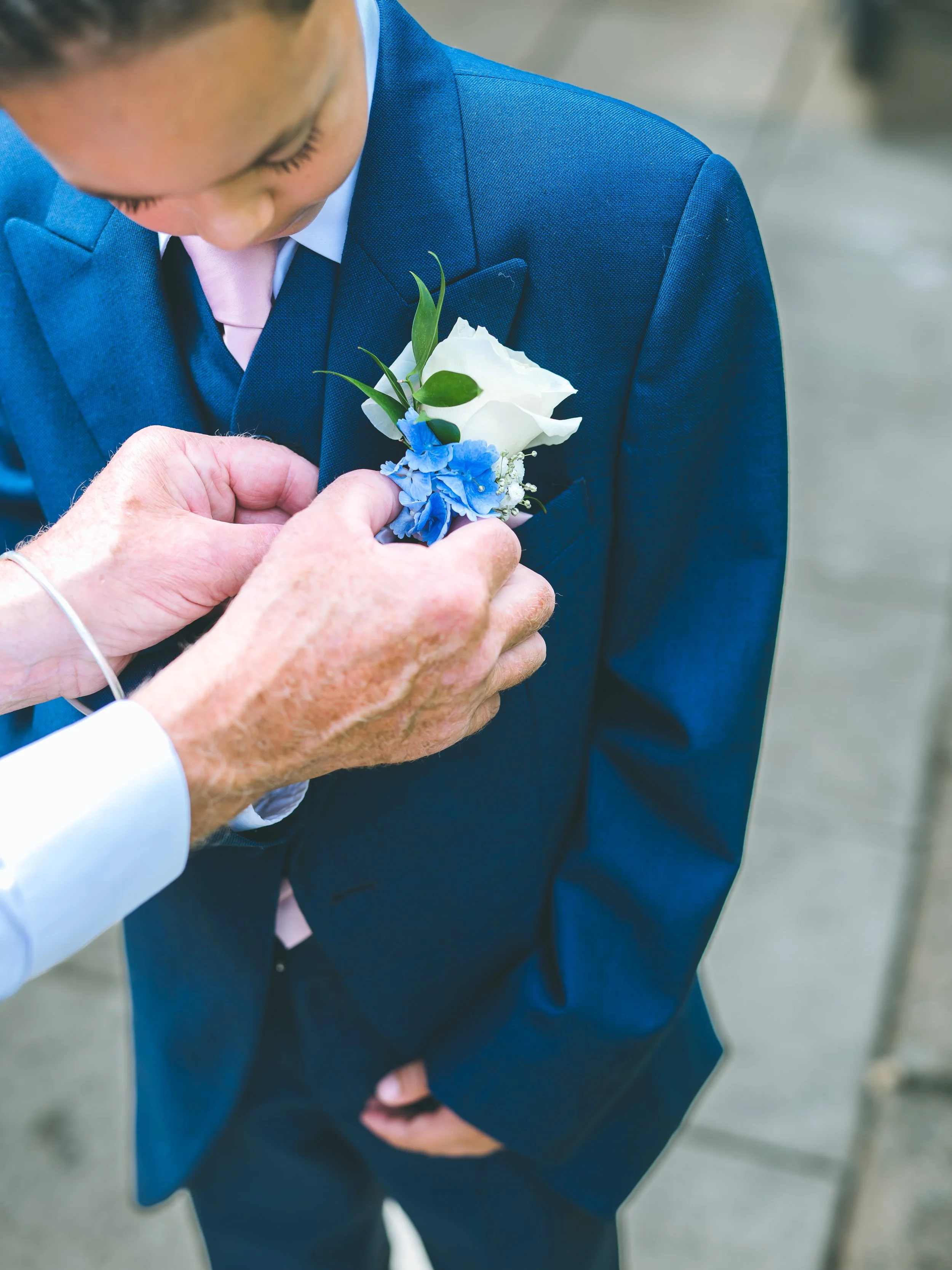 A man in a blue suit with a pink tie is pinning a white rose and blue flowers on a woman's dress during a formal event or wedding.