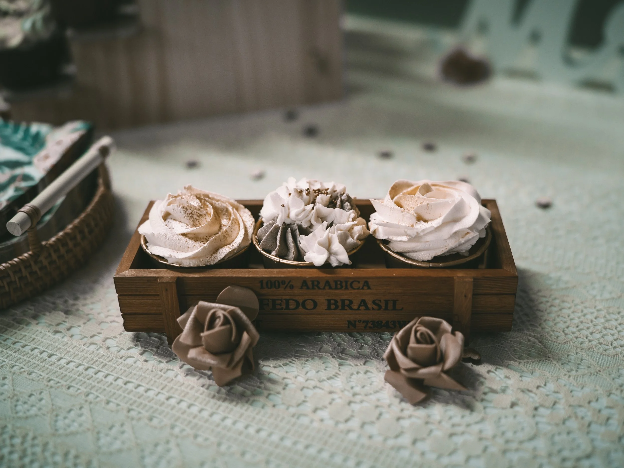 A wooden tray with coffee cups decorated with whipped cream and sprinkles, placed on a lace tablecloth with a beige fabric rose decoration nearby.