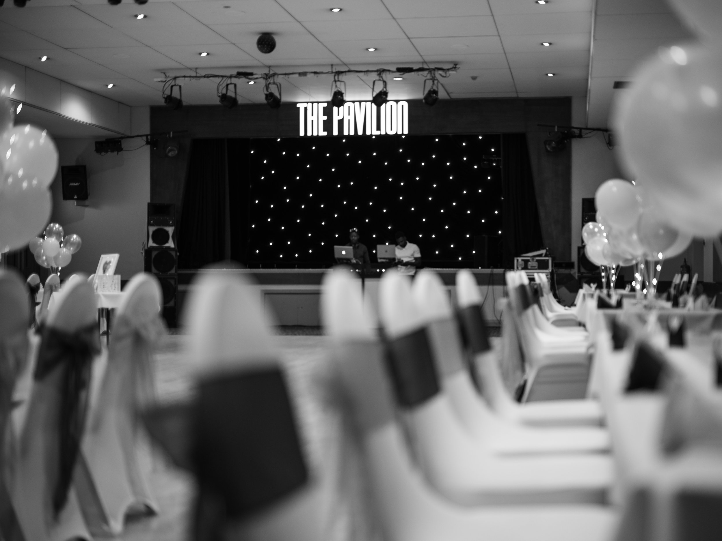 A stage with a sign that reads 'The Pavilion' and two DJs working at their equipment, surrounded by balloons and tables in the foreground.