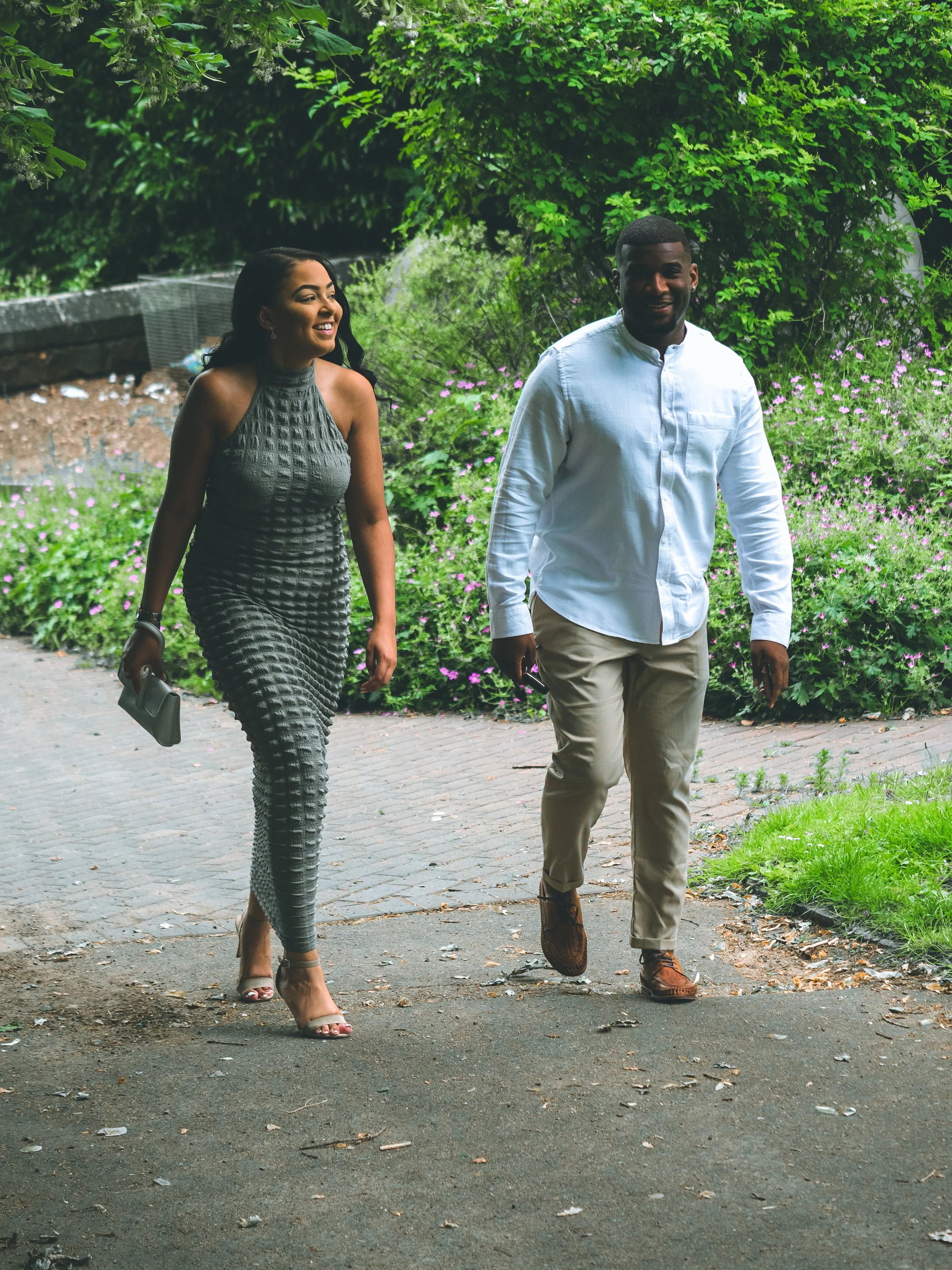A woman in a sleeveless gray dress and heels walking and smiling next to a man in a white shirt and beige pants, also smiling, walking outdoors on a paved pathway with green trees and pink flowers in the background.