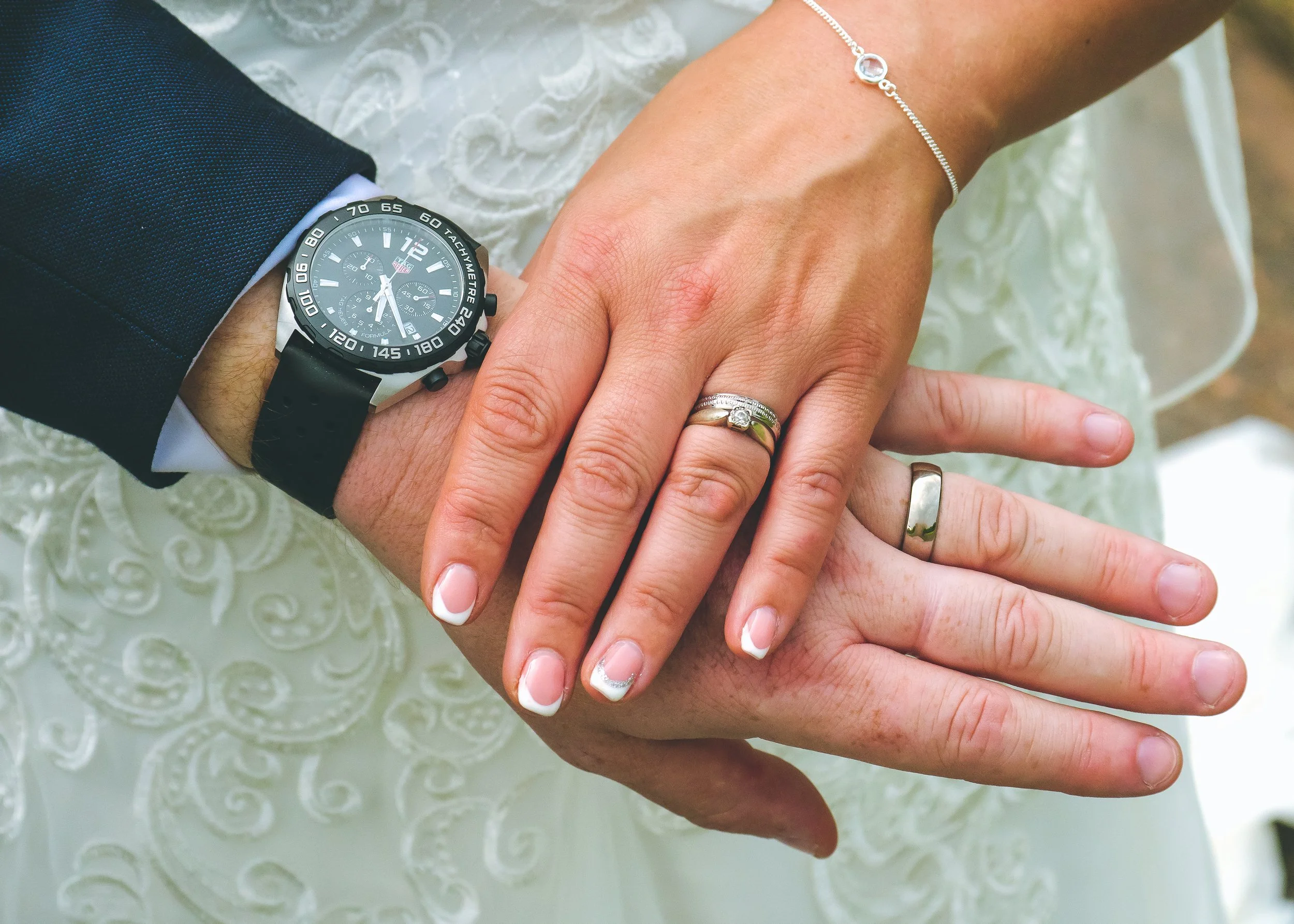 Close-up of a couple's hands intertwined, showing wedding rings and a wristwatch, against a white lace fabric background.