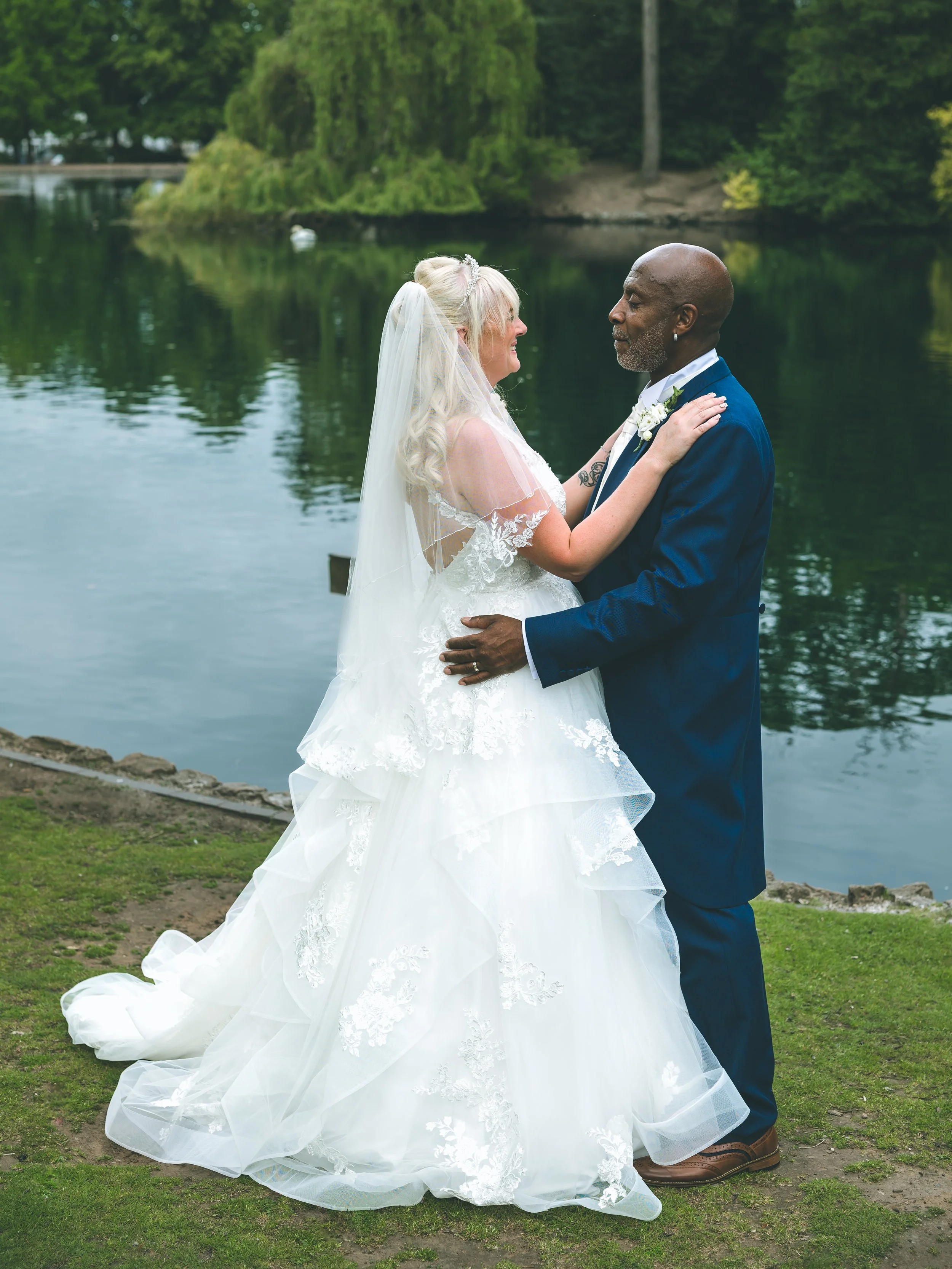 A bride and groom standing close by a lake, looking at each other, with trees and water in the background.