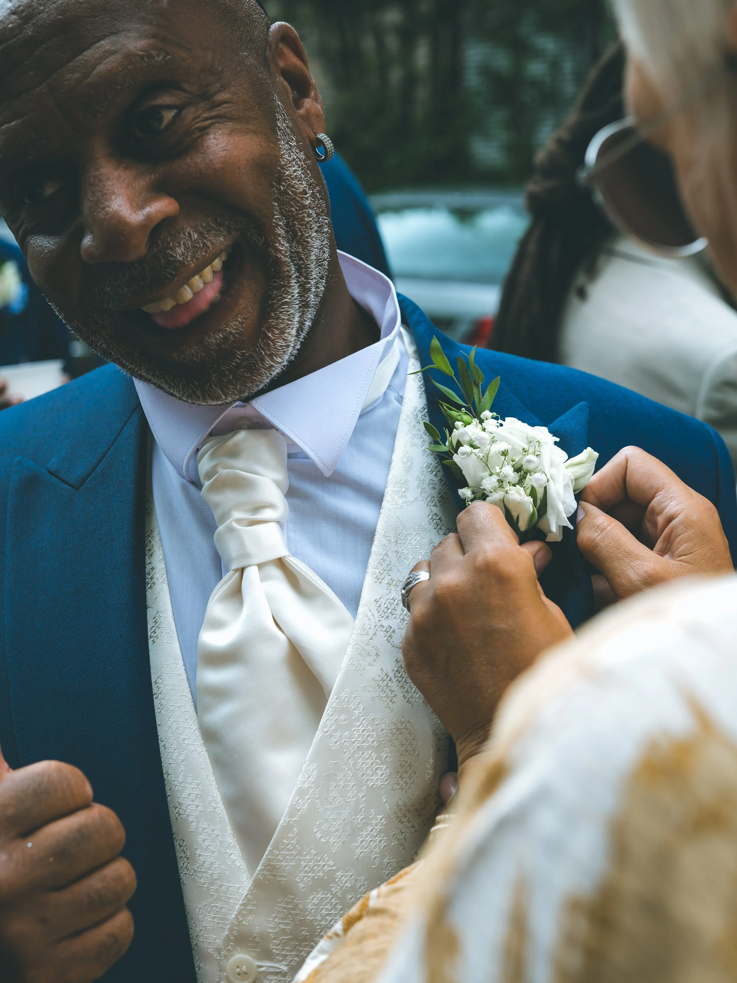 A man with gray hair and a beard smiling, wearing a blue suit and white shirt, pinning a white flower boutonniere on a person's clothing during a celebration or event.