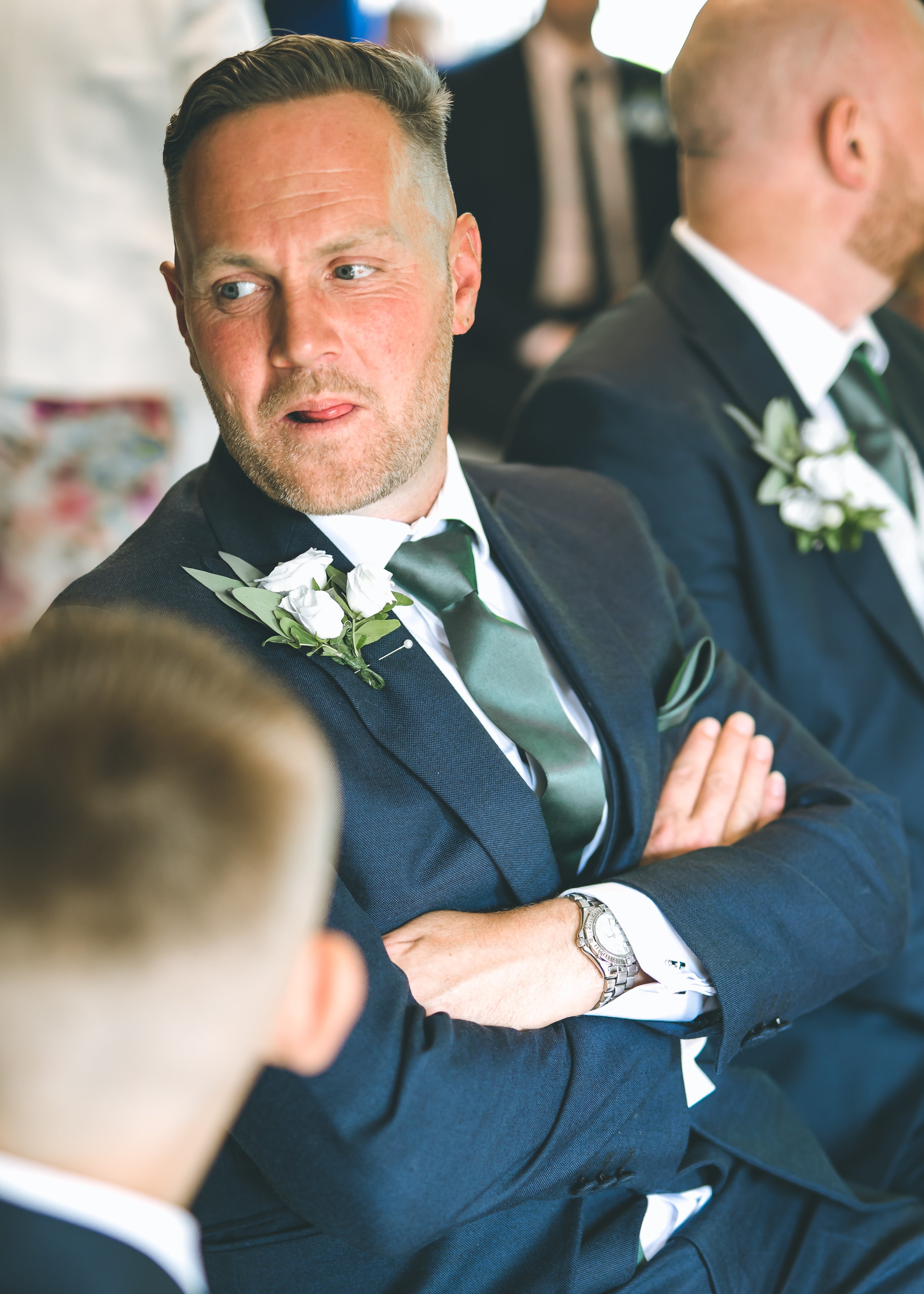 A man with light skin, styled hair, and a beard, dressed in a dark suit with a white shirt, a black tie, and a boutonniere, is sitting with his arms crossed, looking to his left. Another man dressed similarly is next to him, partially visible.