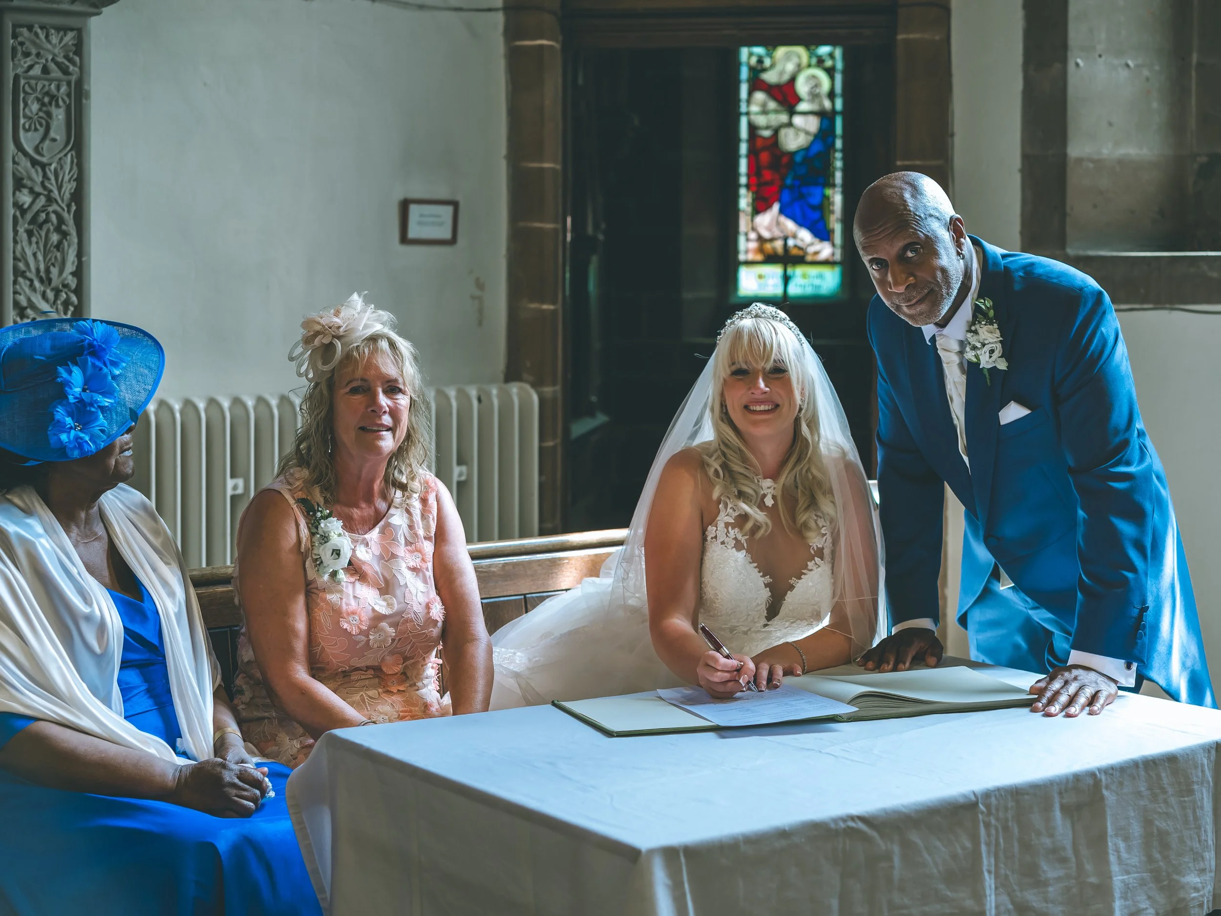 A wedding ceremony inside a church with five people gathered around a table. The bride, a blonde woman in a wedding gown, is sitting and smiling as she signs a document. To her right, a man in a blue tuxedo is leaning over the table, holding her hand