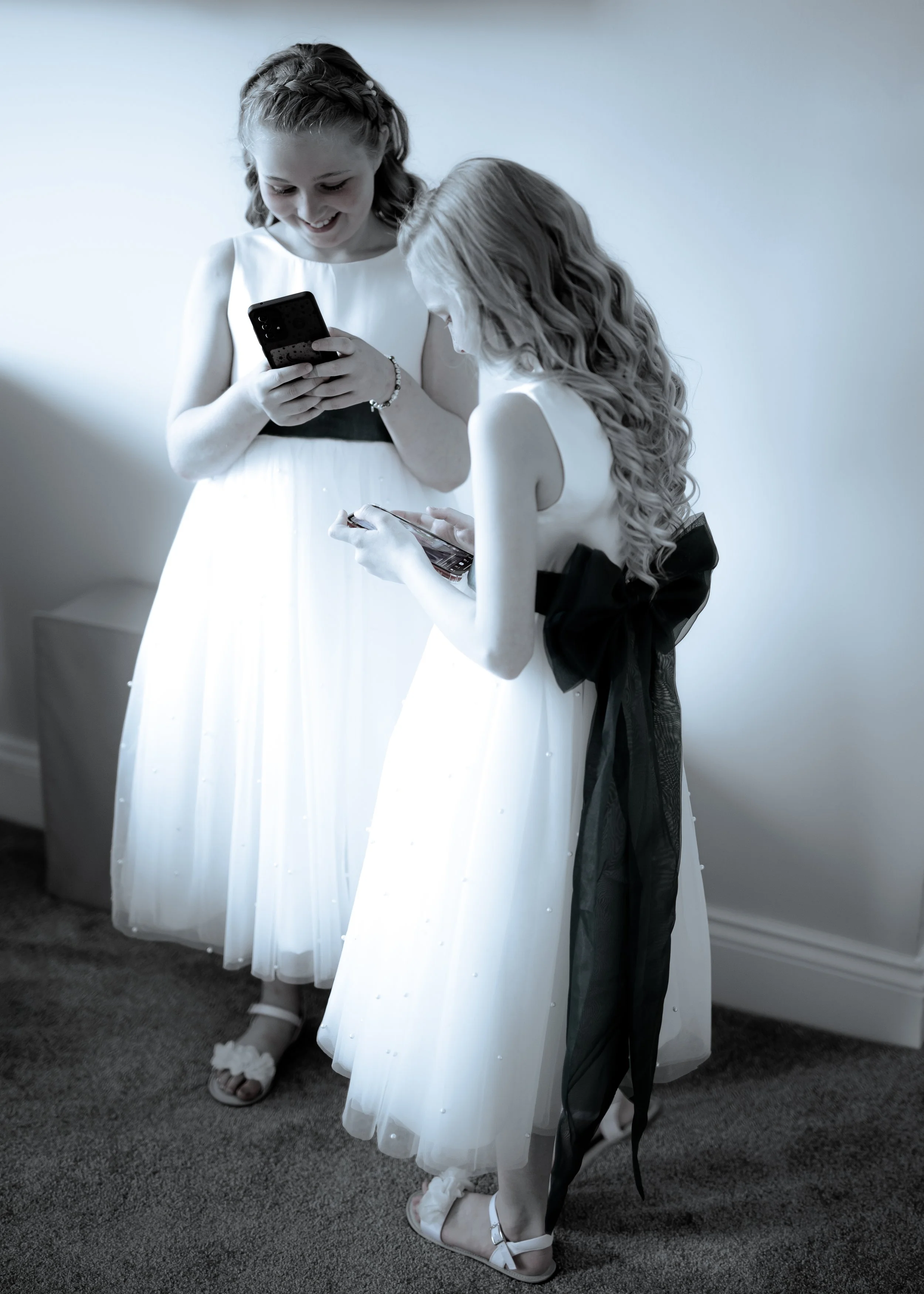 Two girls in white dresses look at their phones, standing on a gray carpeted floor against a plain wall.
