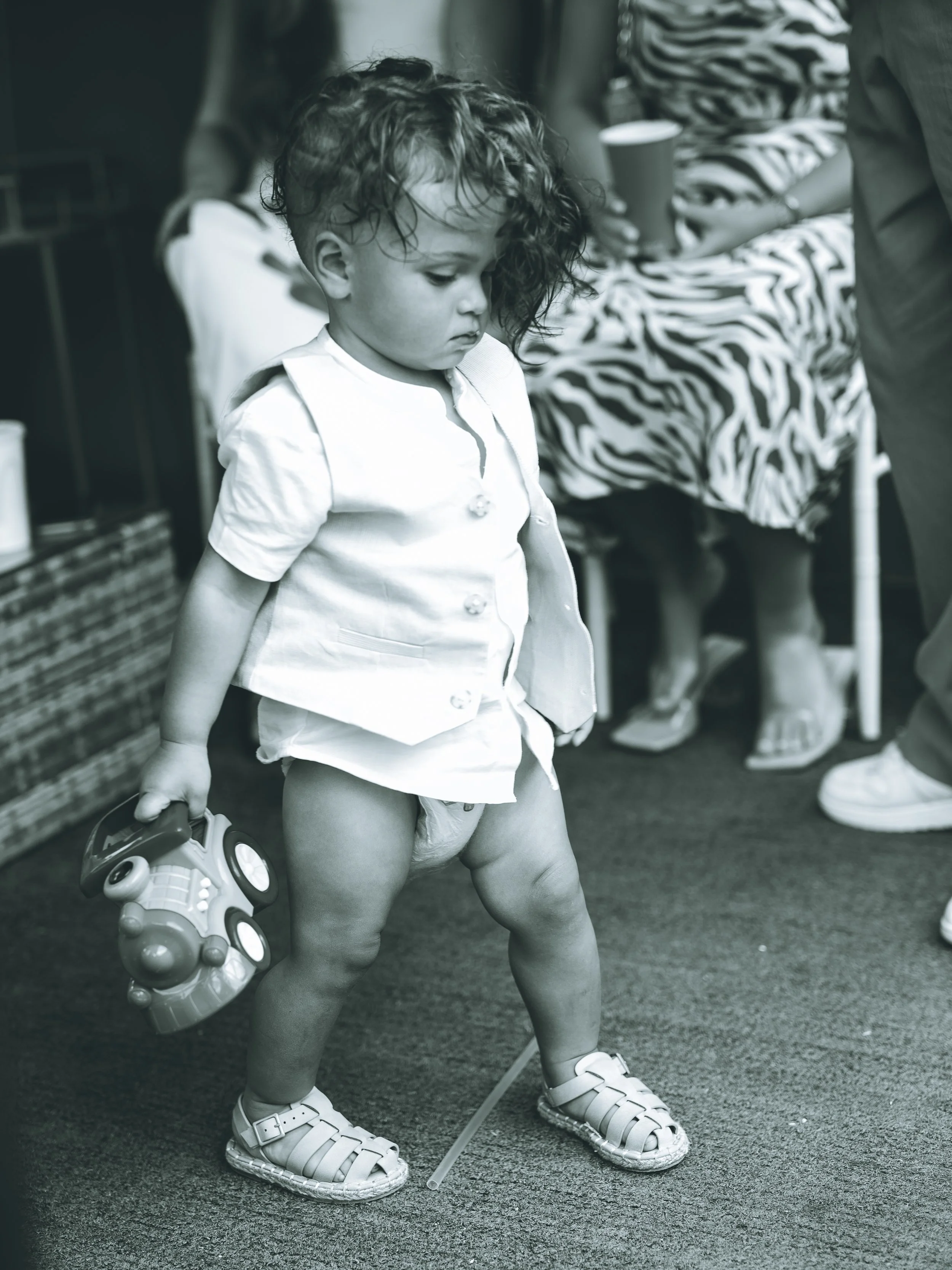 A young child with curly hair dressed in a white vest and shorts, wearing large white sandals, holding a toy vacuum cleaner in one hand, standing on a carpeted floor in an indoor setting with people in the background.