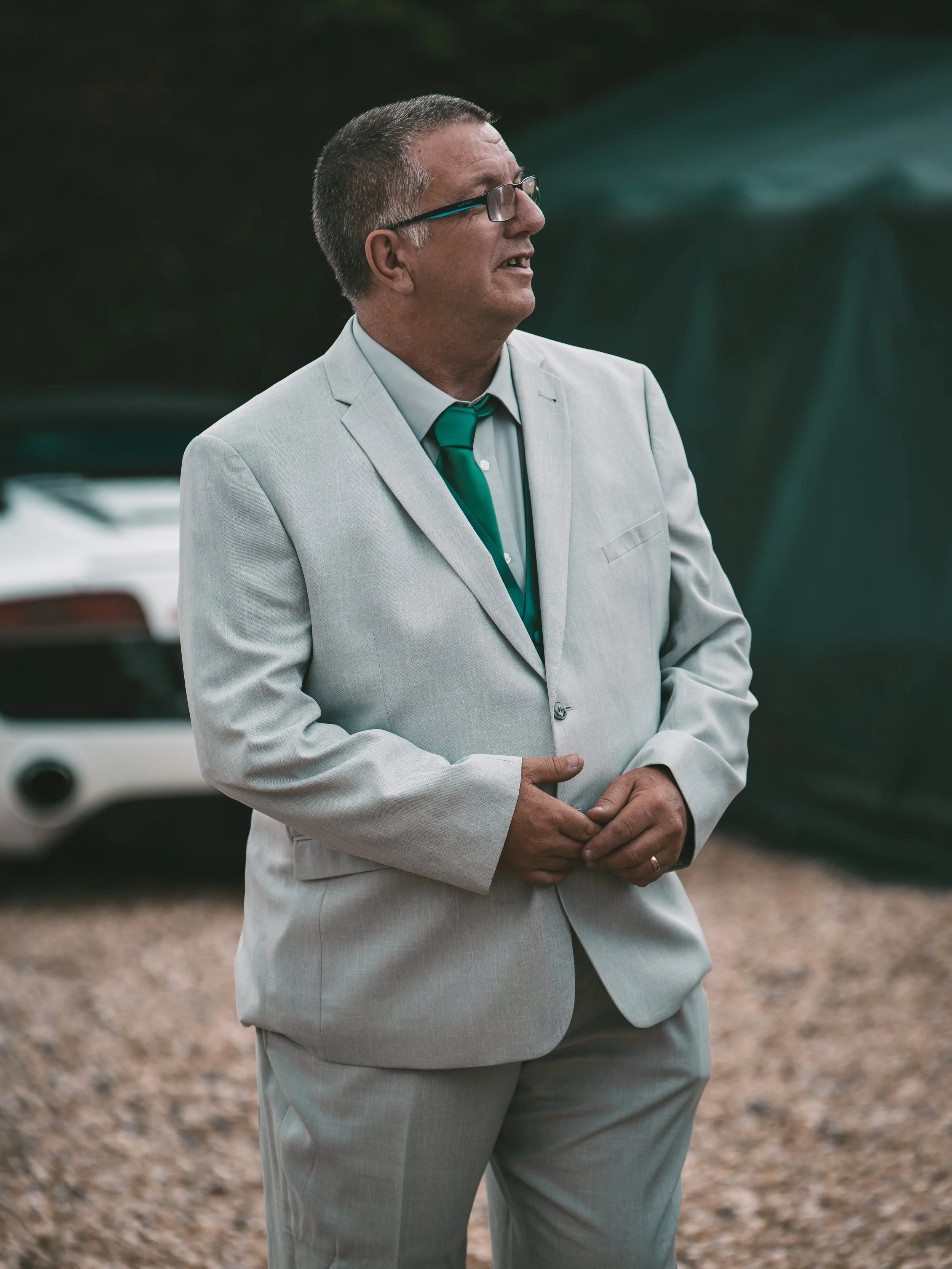 A man in a light gray suit with a teal tie standing outdoors with other cars in the background.