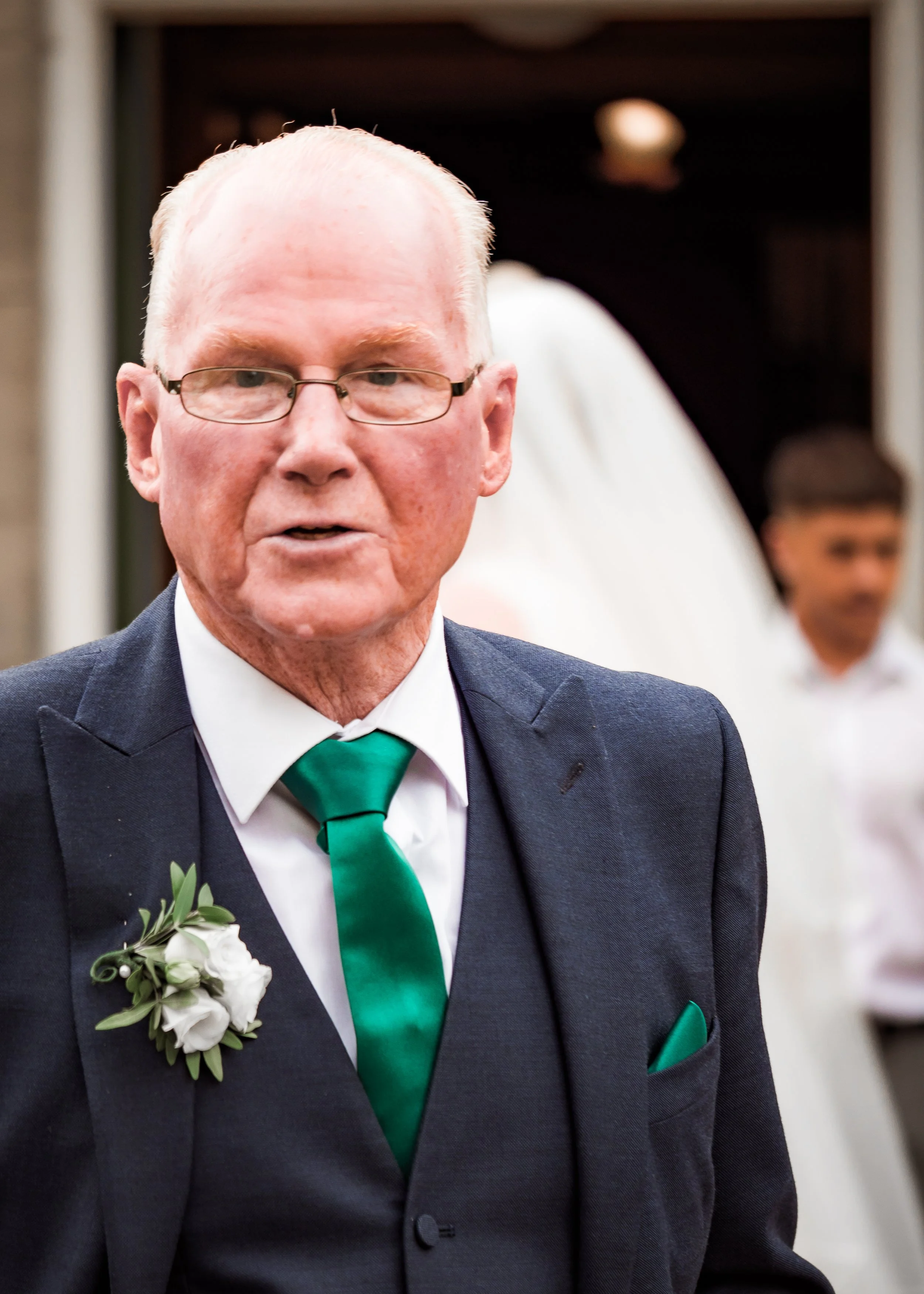 Close-up of a man in a dark suit with a green tie, white shirt, and boutonnière, attending a formal event or celebration.