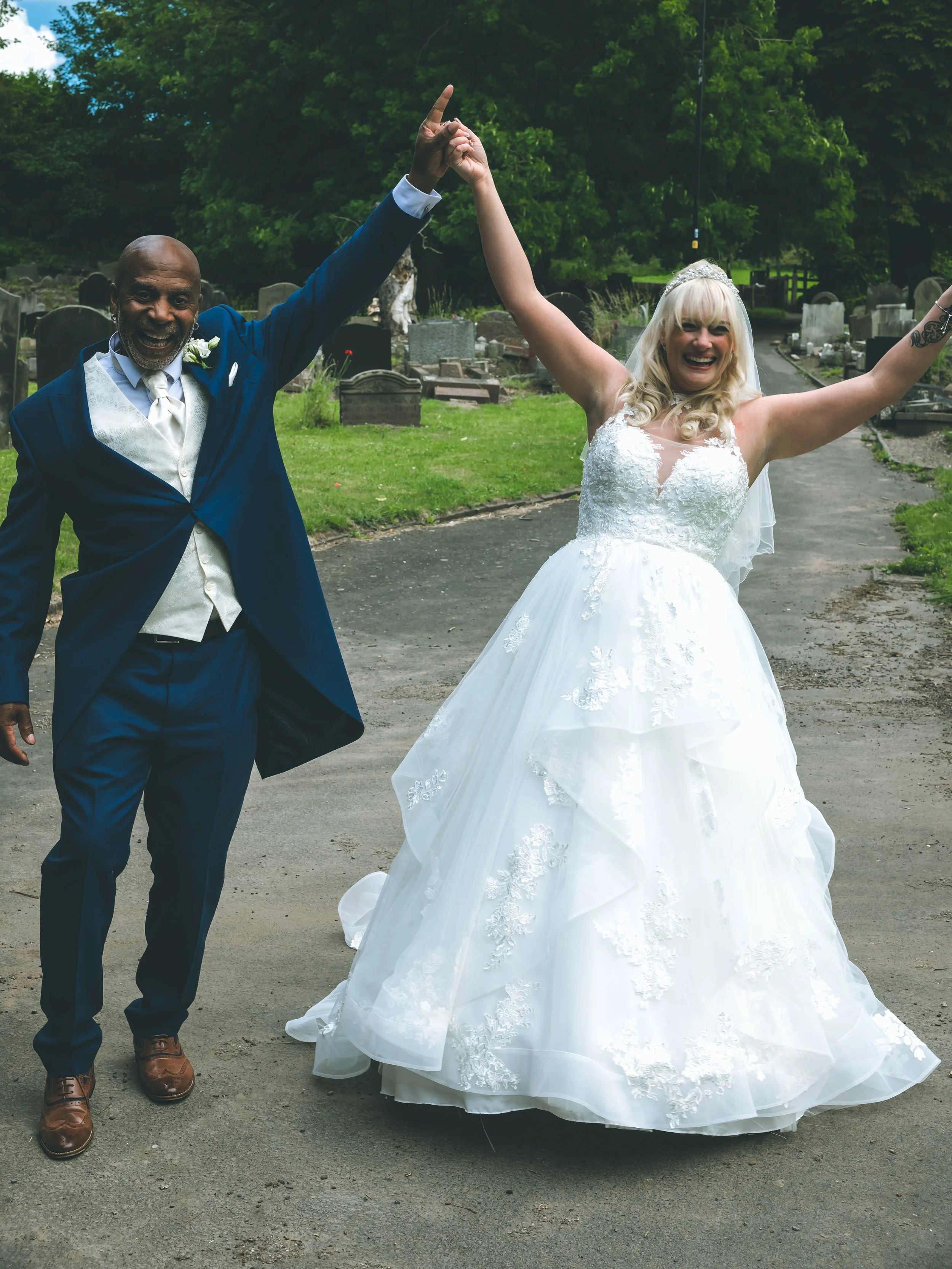 A joyful couple in wedding attire holding hands and celebrating outdoors in a cemetery with green trees and tombstones.