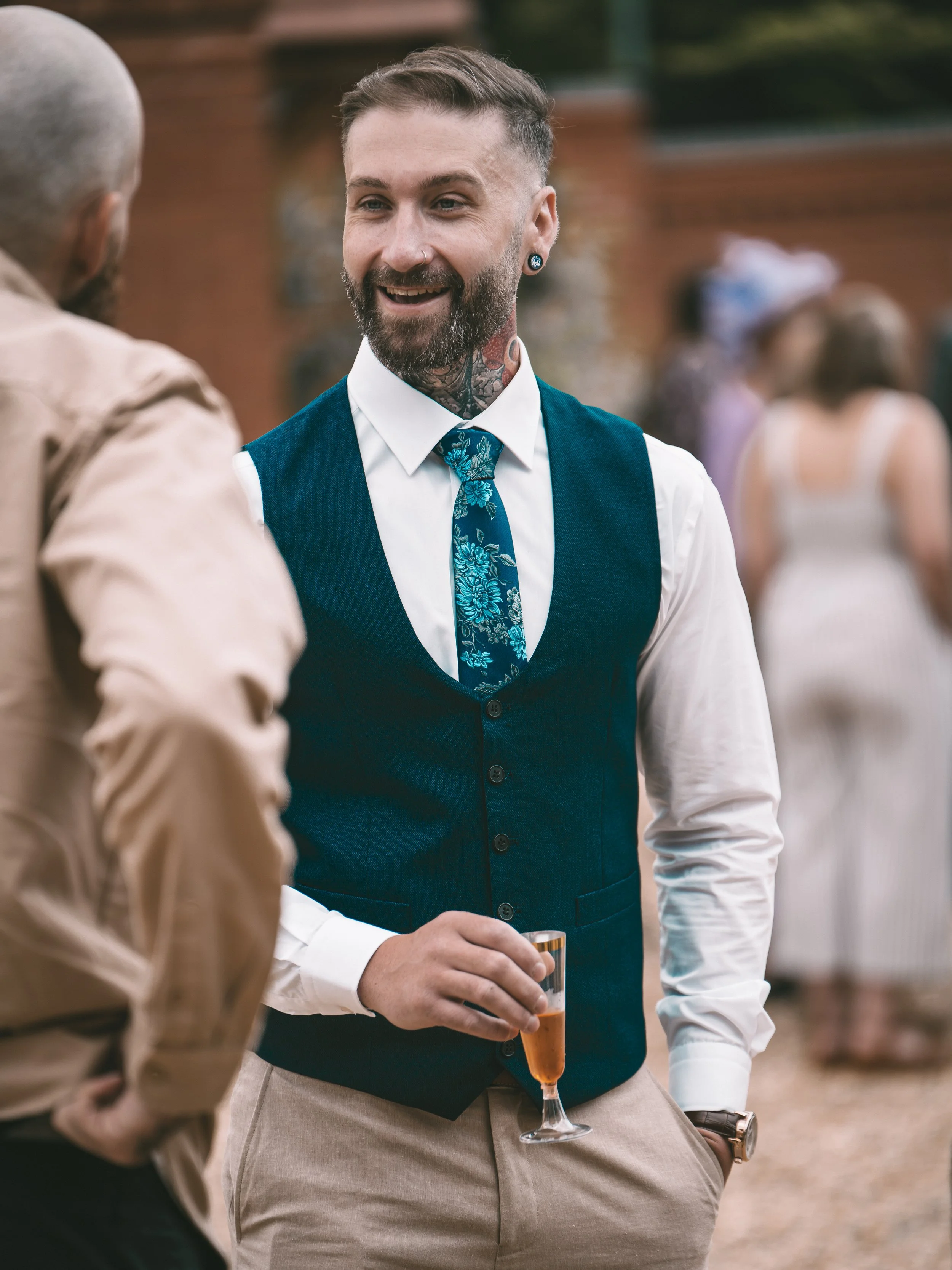 A man with tattoos, a beard, and earrings is holding a glass of drink and smiling while talking to another person at an outdoor event.