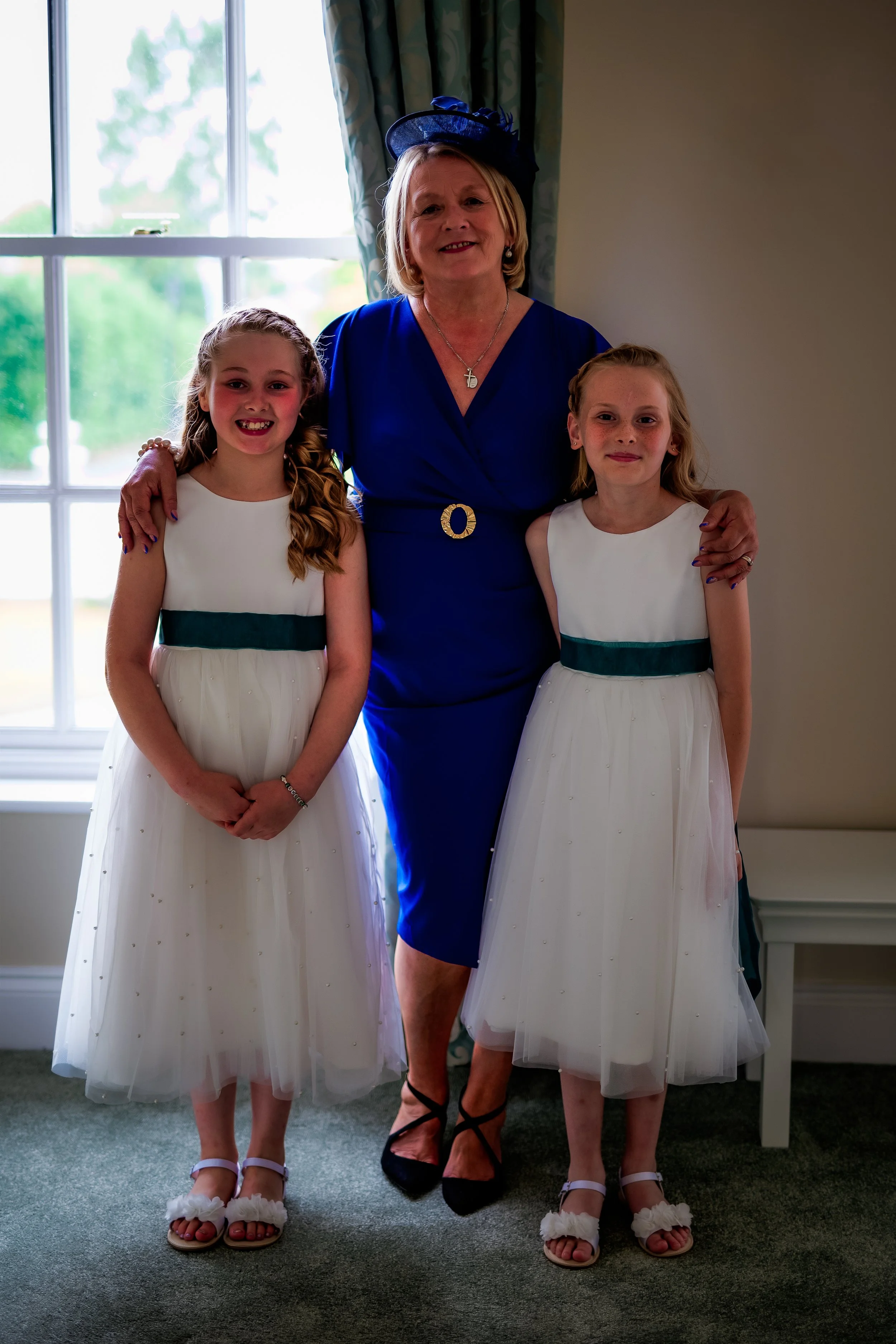 A group of three females standing inside a room with a window in the background. Two young girls are wearing white dresses with dark green sashes, and an older woman in a blue dress and a matching hat stands between them, smiling.