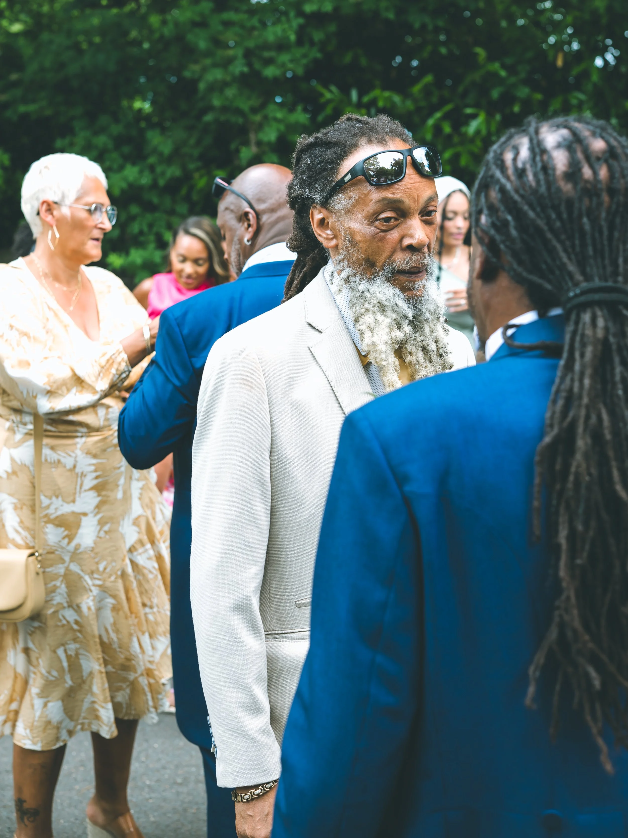 Group of diverse people outdoors in conversation, focus on older man with beard and sunglasses, others in background include woman with white hair, man in glasses, and young woman with braids.