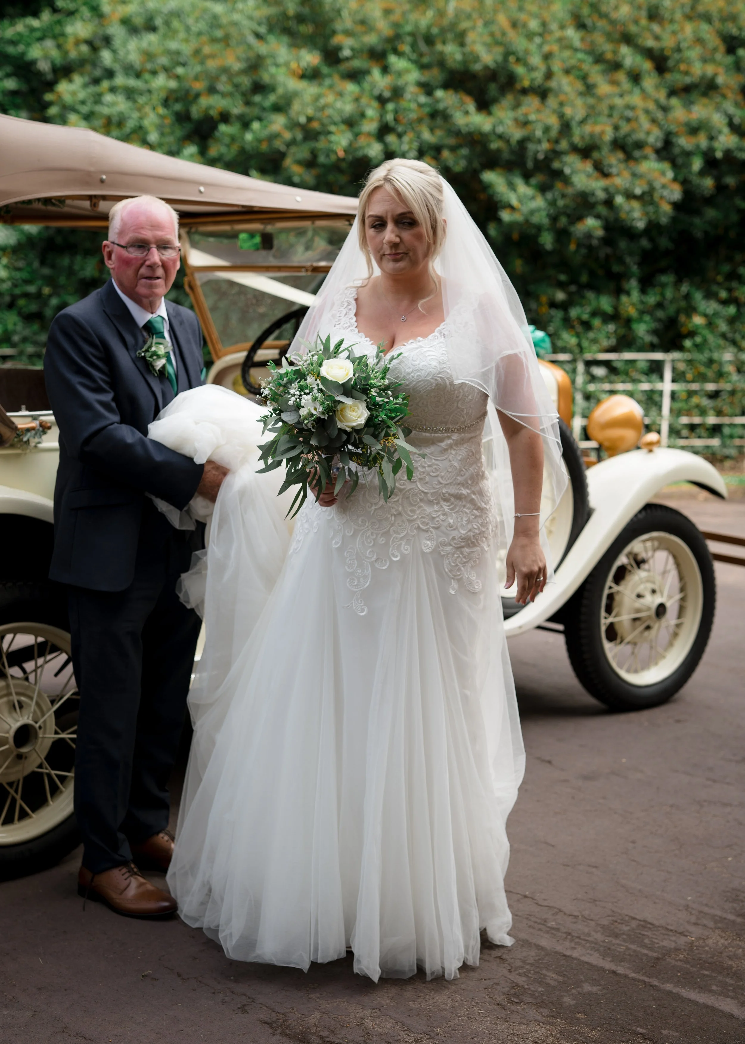 A bride holding a bouquet of white roses and greenery standing beside an older man next to a vintage car outdoors with green trees in the background.
