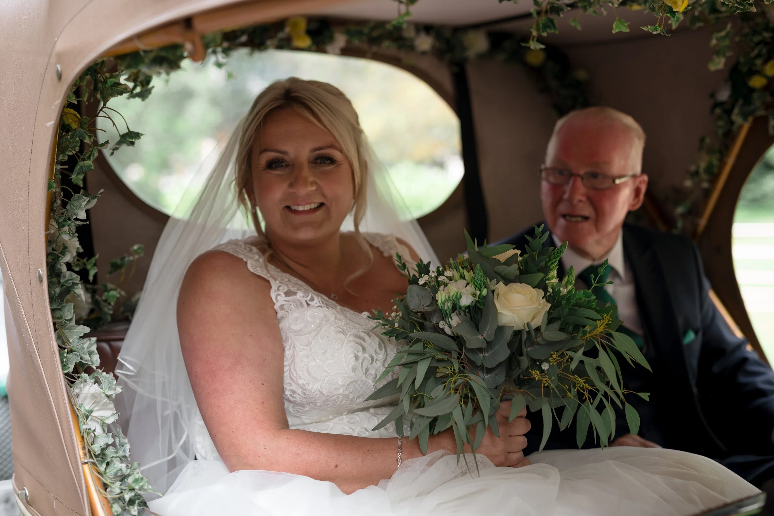 A bride with blonde hair smiling holding a bouquet of white and green flowers inside a decorated carriage with ivy and flowers, and an elderly man with glasses sitting beside her.