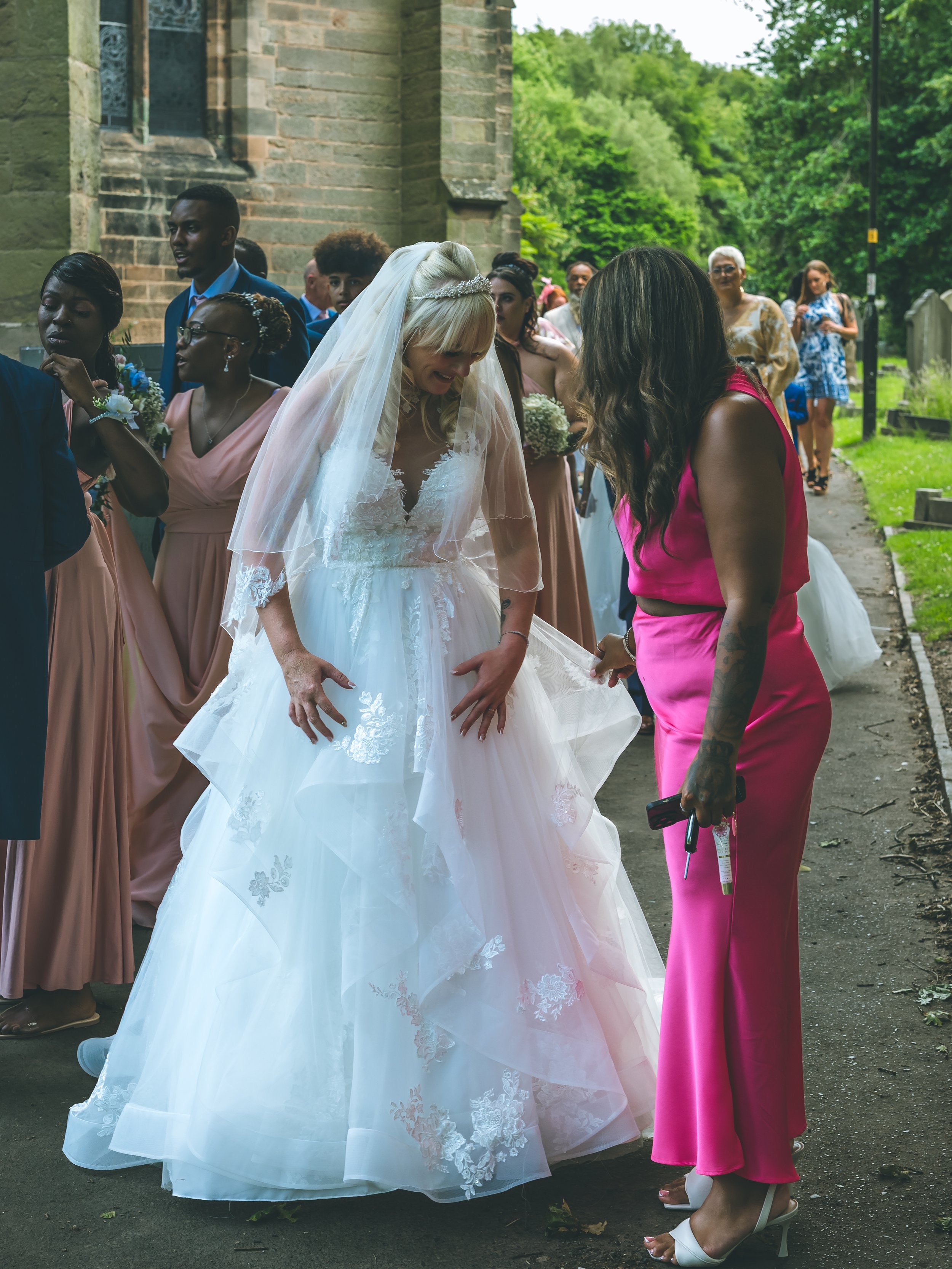 A bride in a white wedding dress and veil is talking to a woman in a pink dress in an outdoor setting near a church or historic building, surrounded by friends and family.