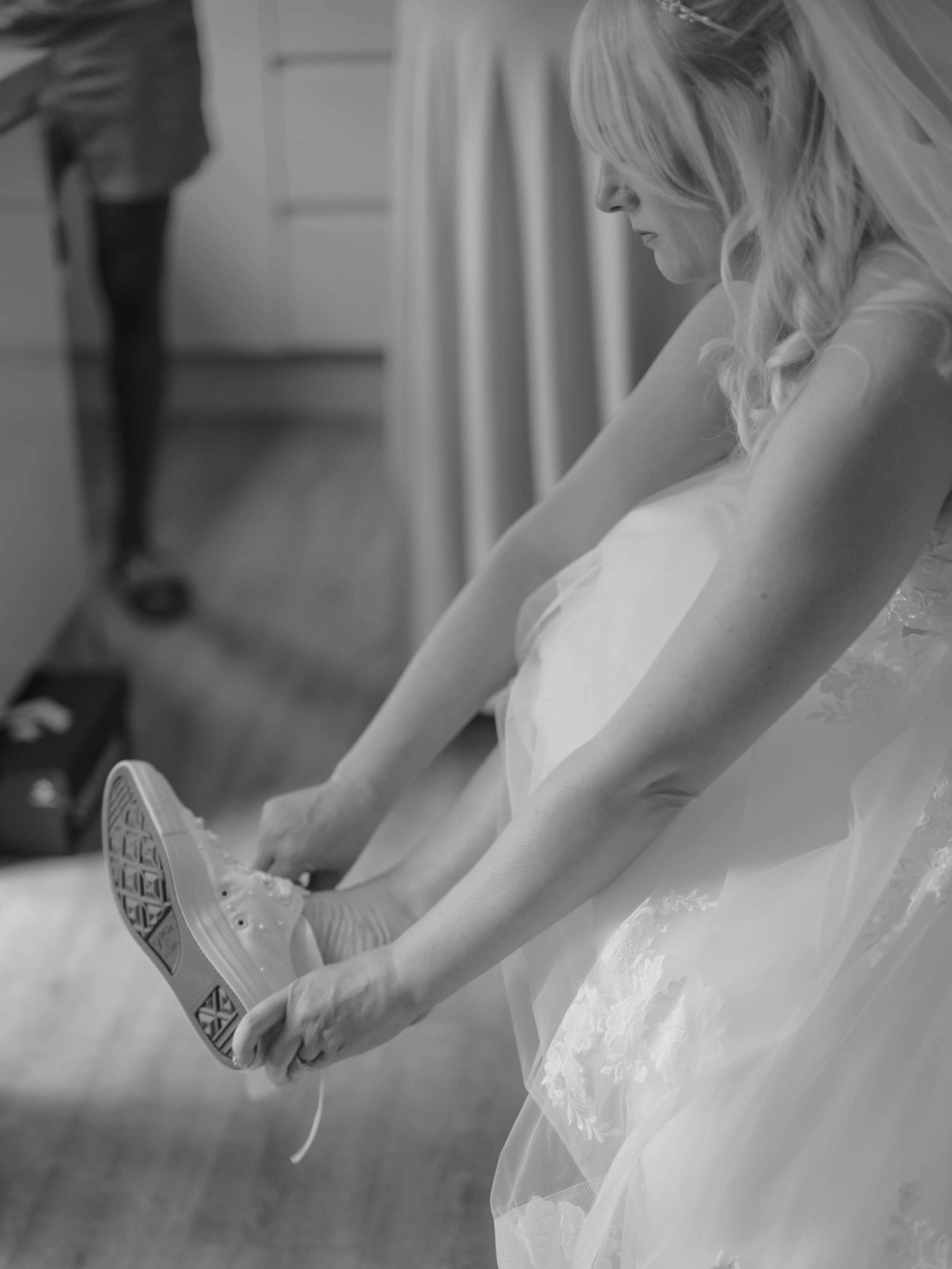 A bride in a wedding dress with lace details sitting on a chair, adjusting her sneakers.