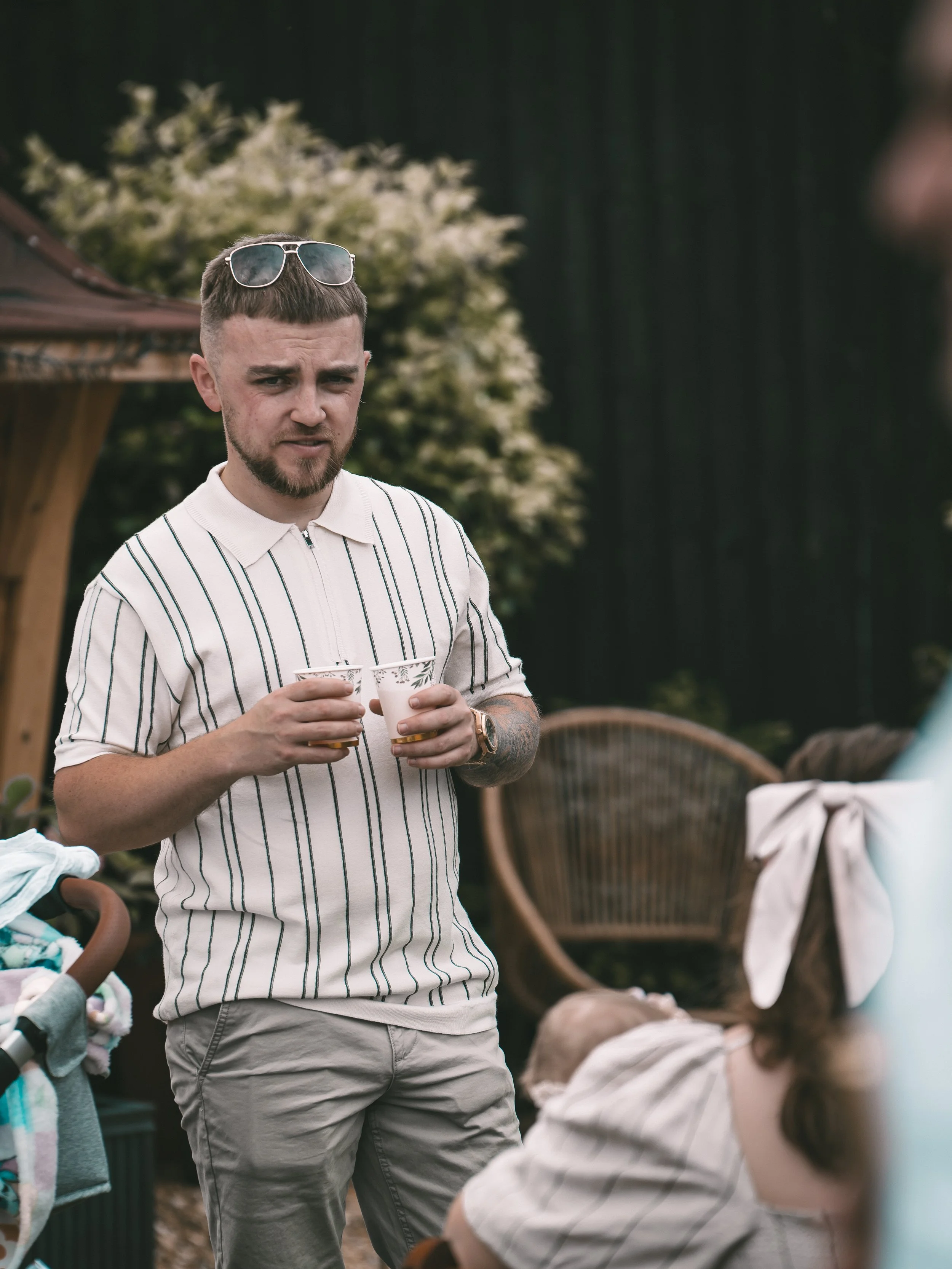 A young man with a beard and short haircut wearing a striped polo shirt, holding two cups, sunglasses resting on his head, standing outdoors among other people at a gathering with a garden and wooden furniture in the background.