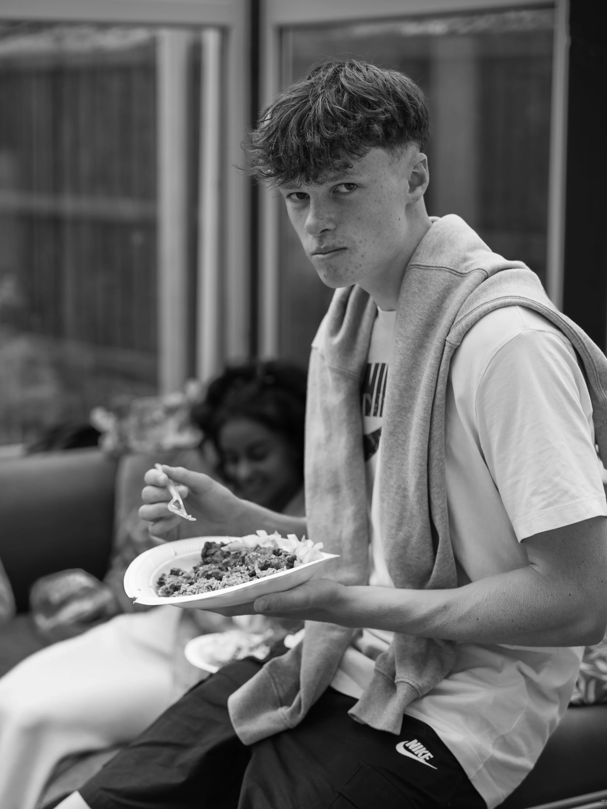 A young man with curly hair and freckles sitting outdoors, eating food from a paper plate.